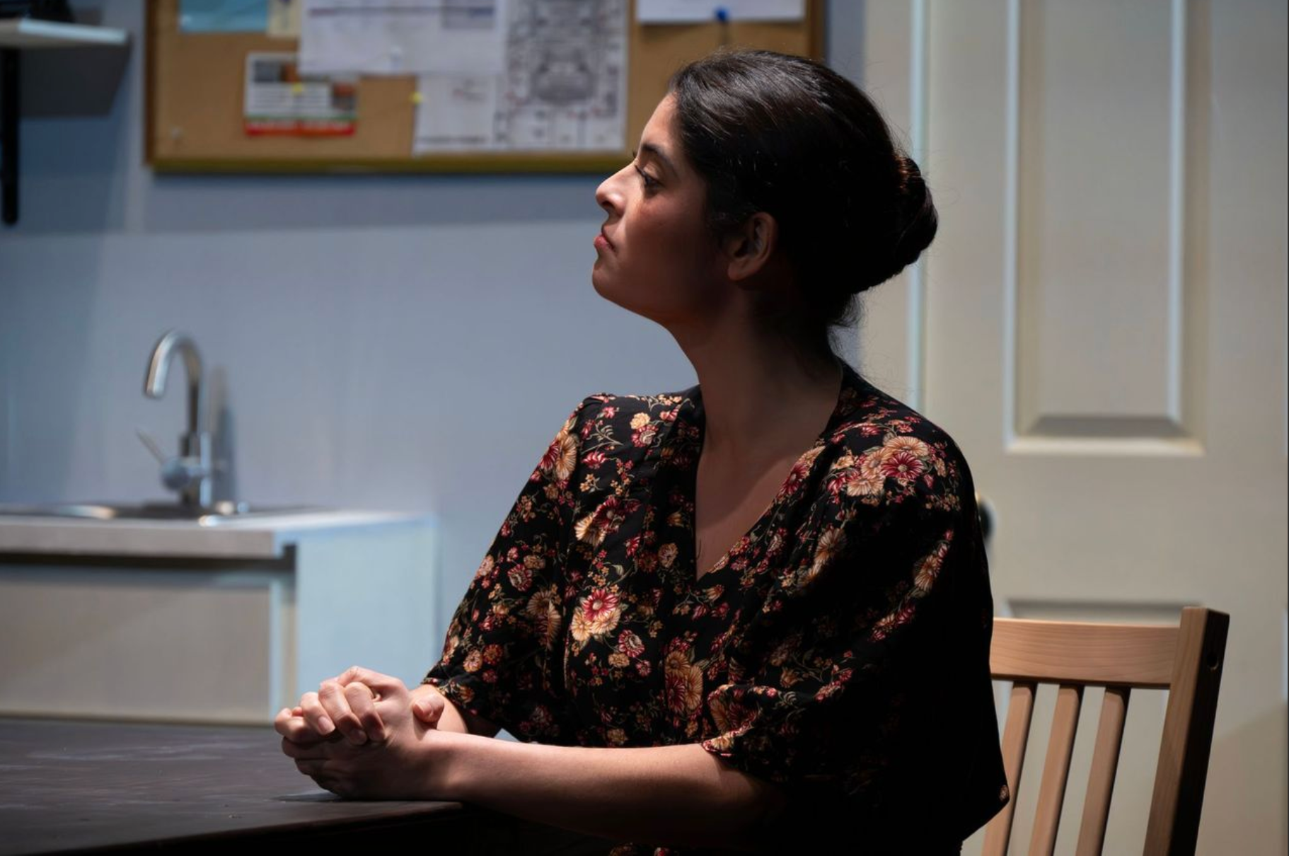 A woman with dark hair styled in a bun is sitting at a table in a room with a bulletin board and a door in the background. She is wearing a black floral blouse and clasping her hands together on the table.