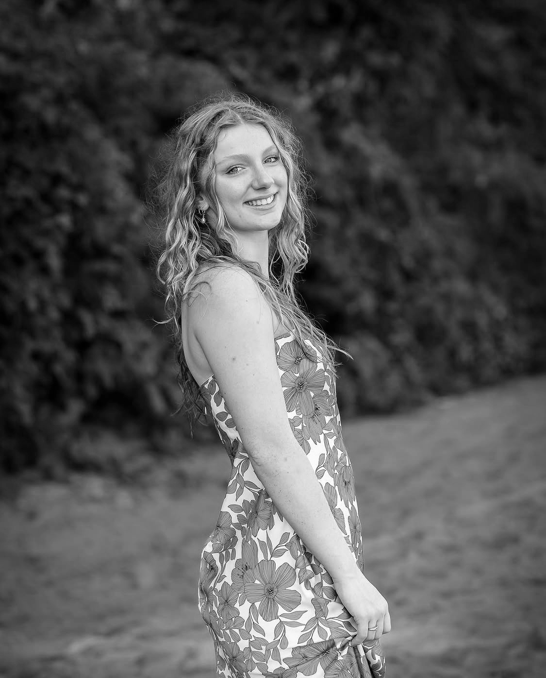 Black-and-white photo of a young woman smiling, wearing a floral dress, standing outdoors against a natural background.