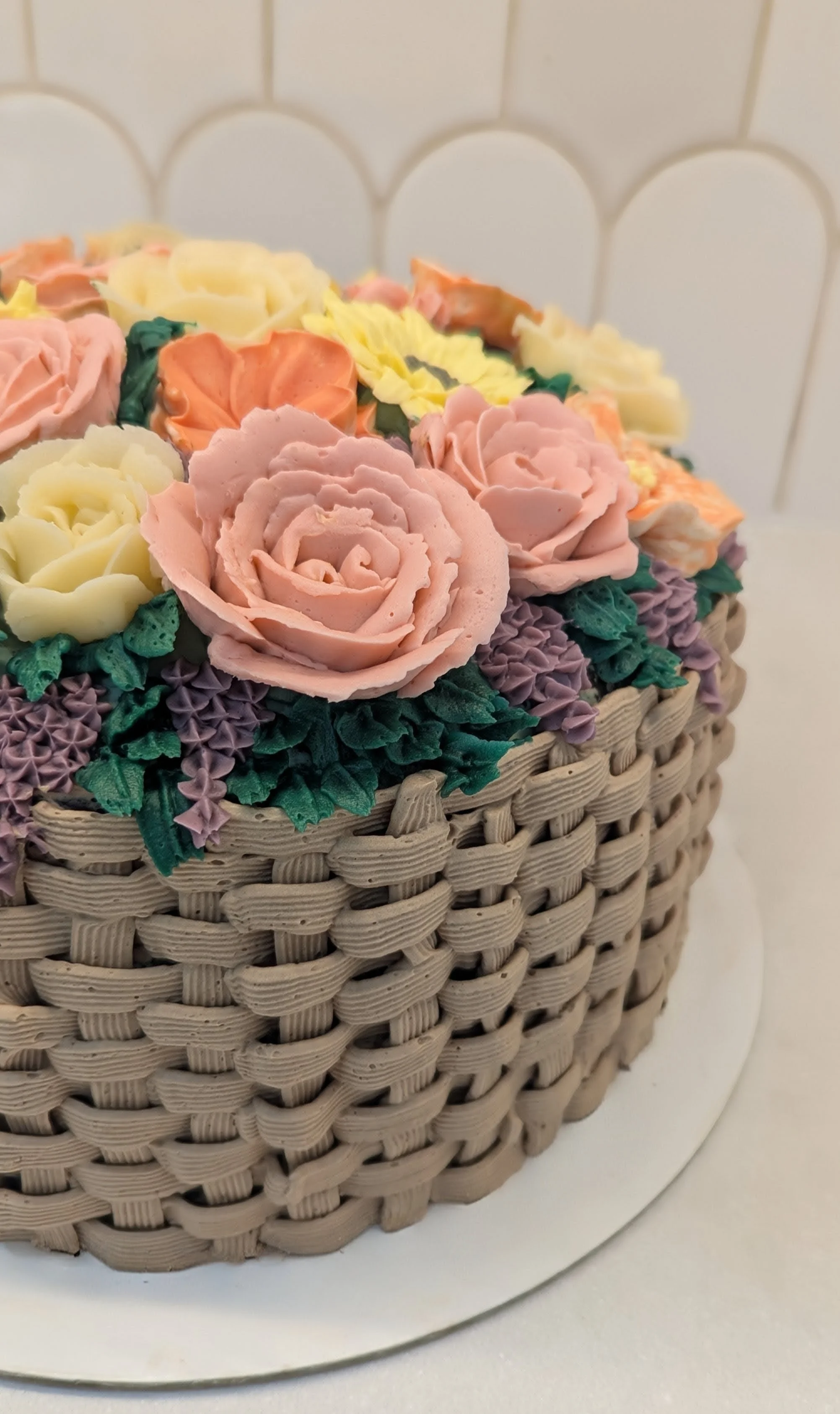 Close-up of a cake decorated with pastel-colored icing flowers and green leaves, with a woven basket-like textured exterior, placed on a white cake board.