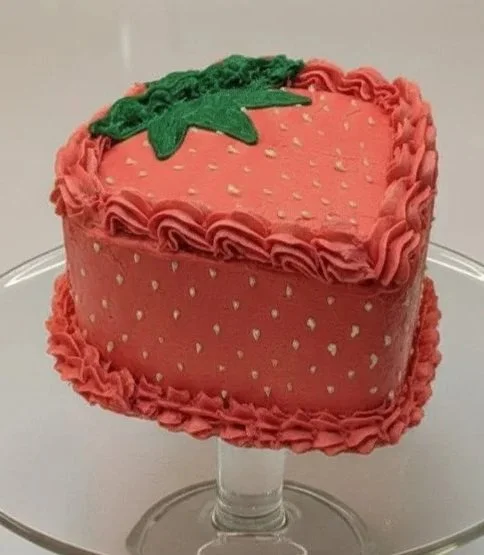 Heart-shaped strawberry-themed cake with pink frosting, white sprinkles, and green leaf decoration on top.