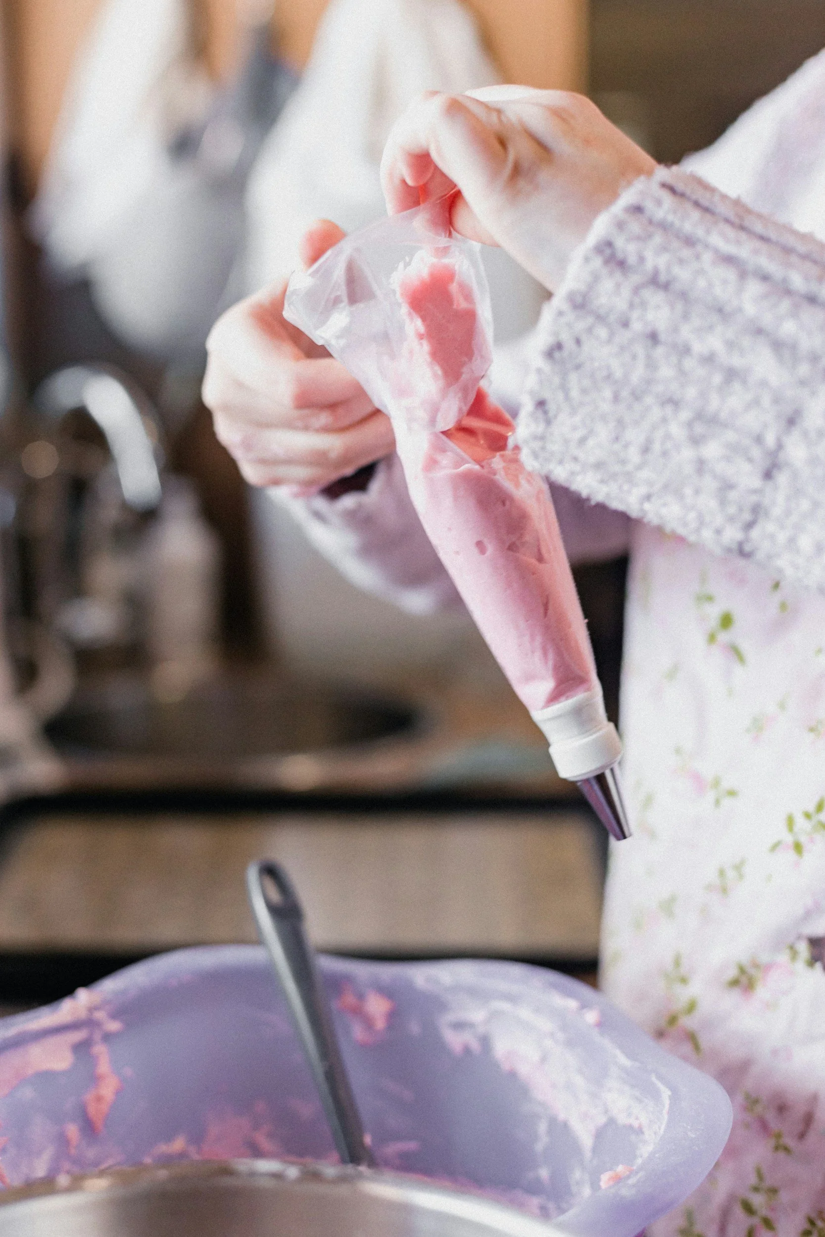 Person holding a piping bag with pink frosting, preparing to decorate a cake. The cake has purple icing with pink decorations and a fork.