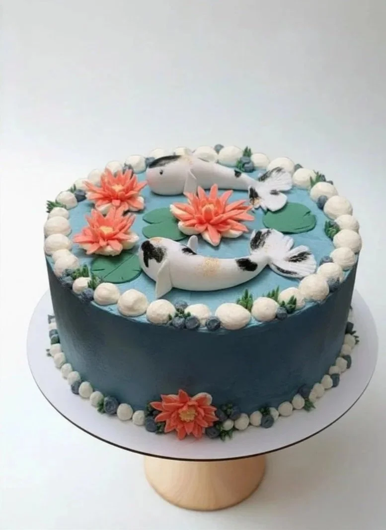 A round cake decorated with white, peach, and pink flowers, green leaves, and two koi fish figurines on top, with a light blue water-themed design, surrounded by white and blue piped borders.