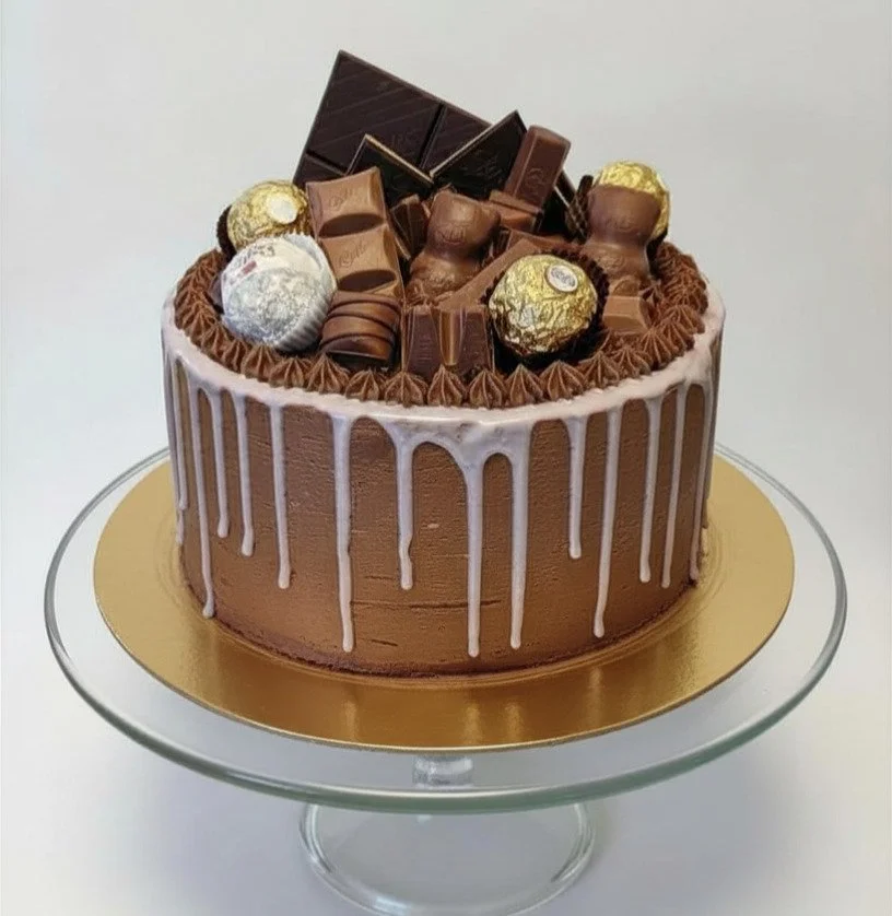 Chocolate cake with gold and silver foil-wrapped chocolates, chocolate bars, and hazelnut-shaped chocolates on top, decorated with white chocolate drips.