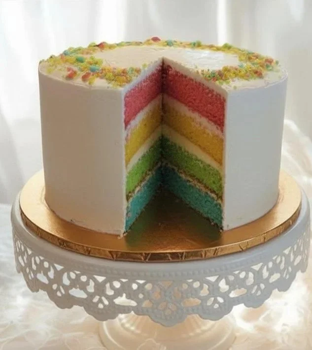A pastel rainbow layered cake with white frosting and colorful sprinkles on top, placed on a decorative white stand.
