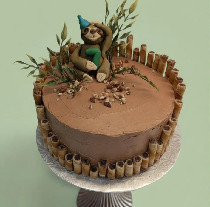 Chocolate cake with a sloth figurine wearing a blue party hat and holding a green number, decorated with wafer rolls around the edge, and green leaves on top.