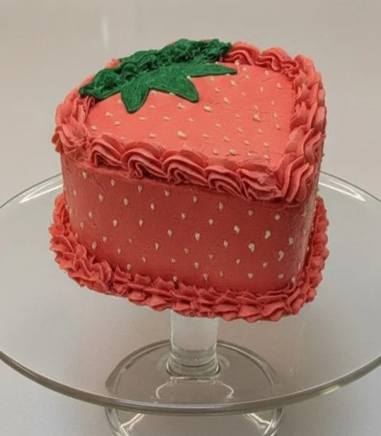 A heart-shaped strawberry-themed cake with pink frosting, white spots, green leaves on top, and decorative pink frosting border, displayed on a clear glass cake stand.