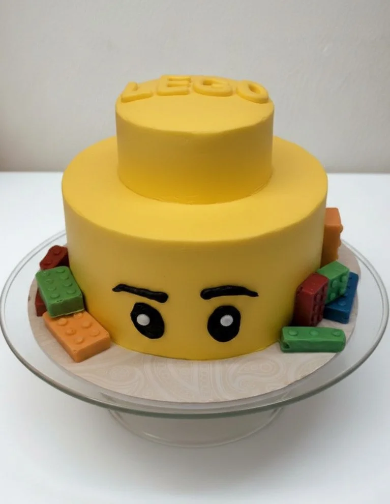 Yellow Lego-themed birthday cake with a face design, surrounded by colorful Lego block decorations, on a clear glass cake stand.