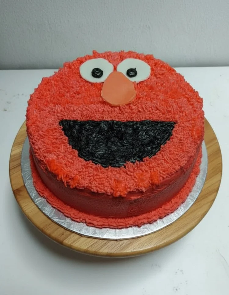Cake decorated to resemble Elmo from Sesame Street, with red frosting, white eyes with black pupils, an orange nose, and a black mouth.