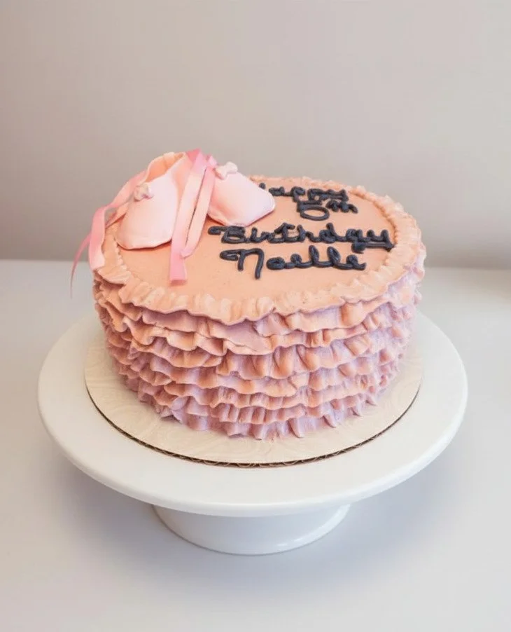 Pink ruffled heart-shaped birthday cake with blue writing and a fondant ballet slipper decoration, on a white cake stand.