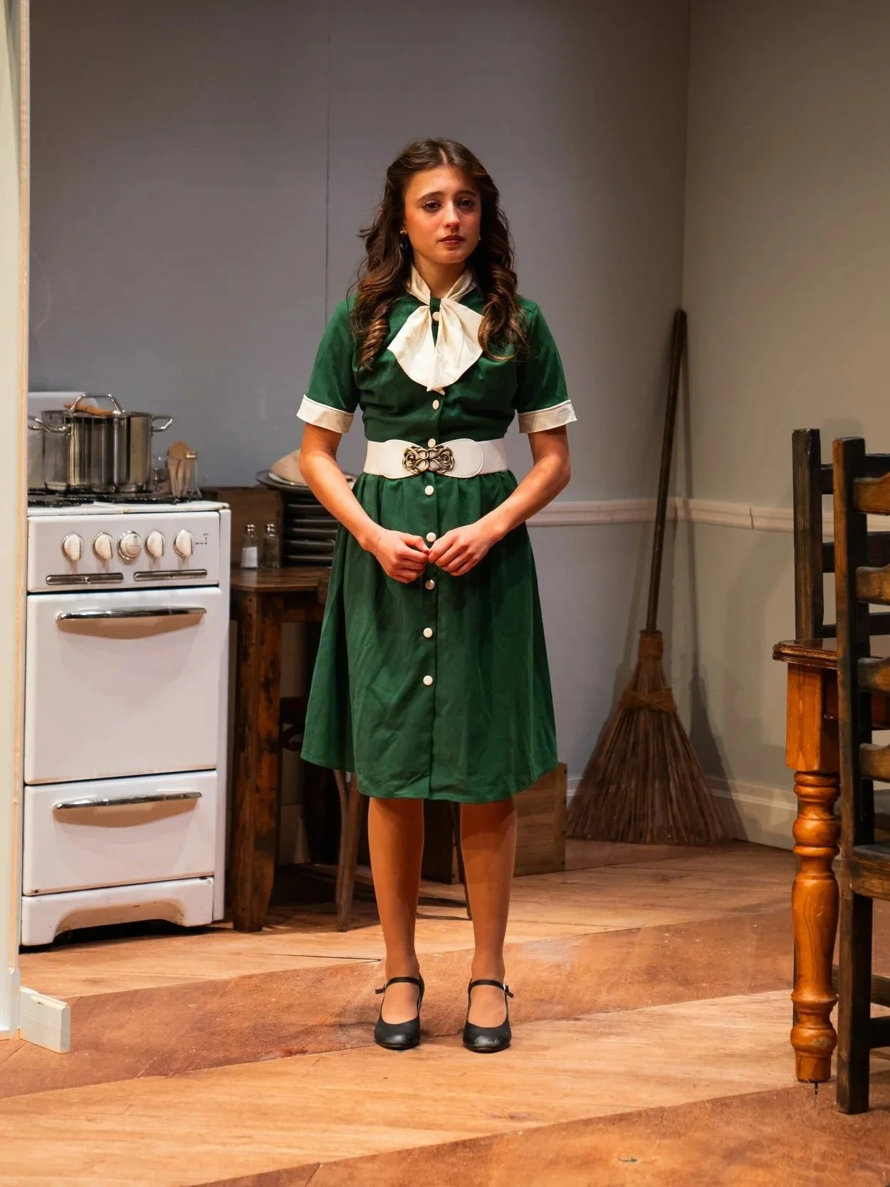 A young woman standing in a kitchen or dining area, wearing a vintage green dress with white accents and a large bow tie, black shoes, and stockings, with a somber facial expression.