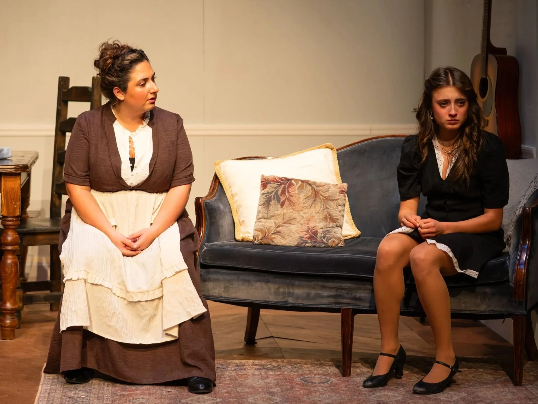 Two women sit in a room with vintage furniture and guitars hanging on the wall. The woman on the left is dressed in period clothing, looking at the woman on the right with a concerned expression. The woman on the right sits with her hands clasped, we