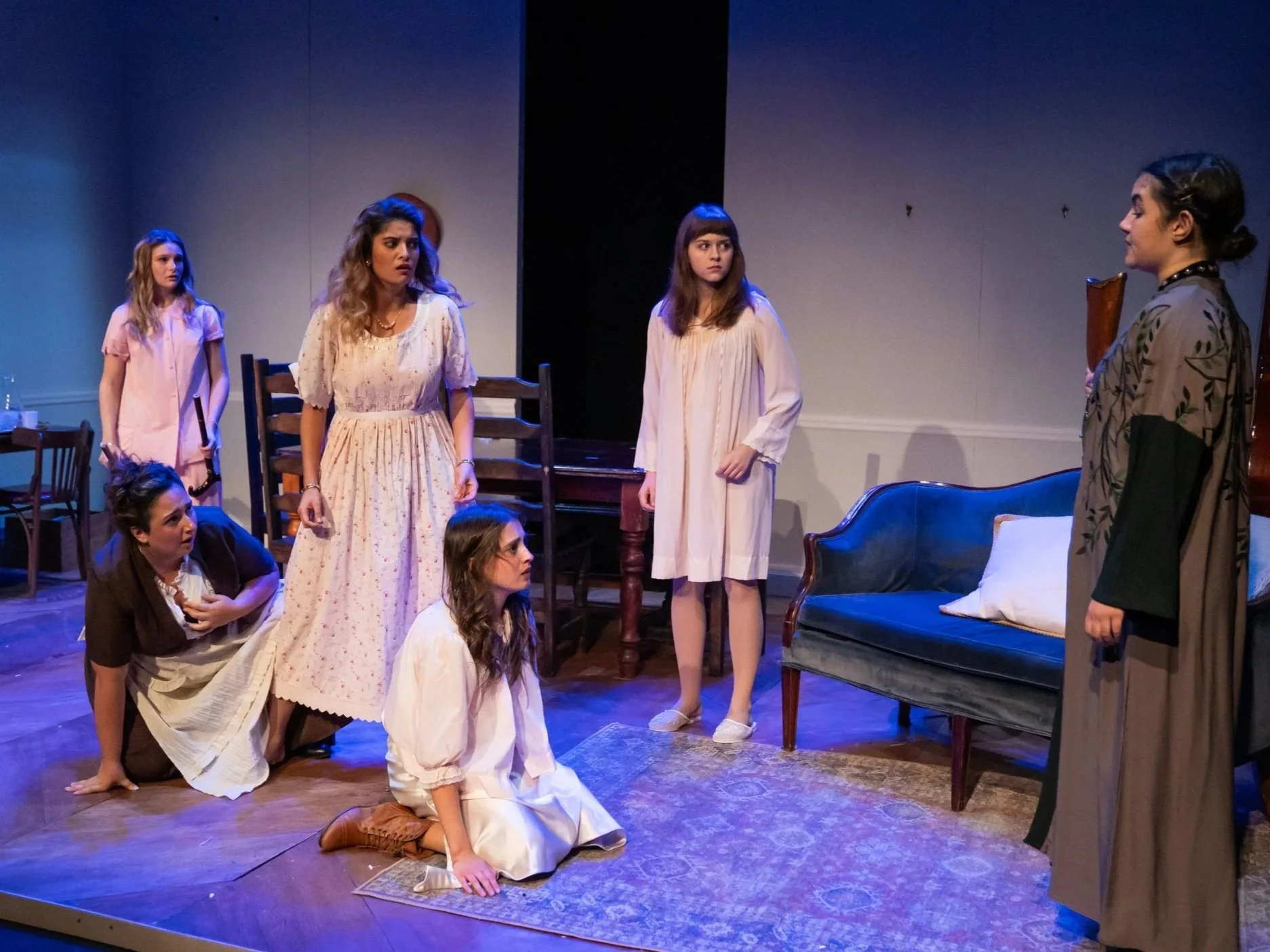 Women in vintage-style clothing on stage performing in a theater play, with some standing and others sitting on the floor, set in a living room scene.