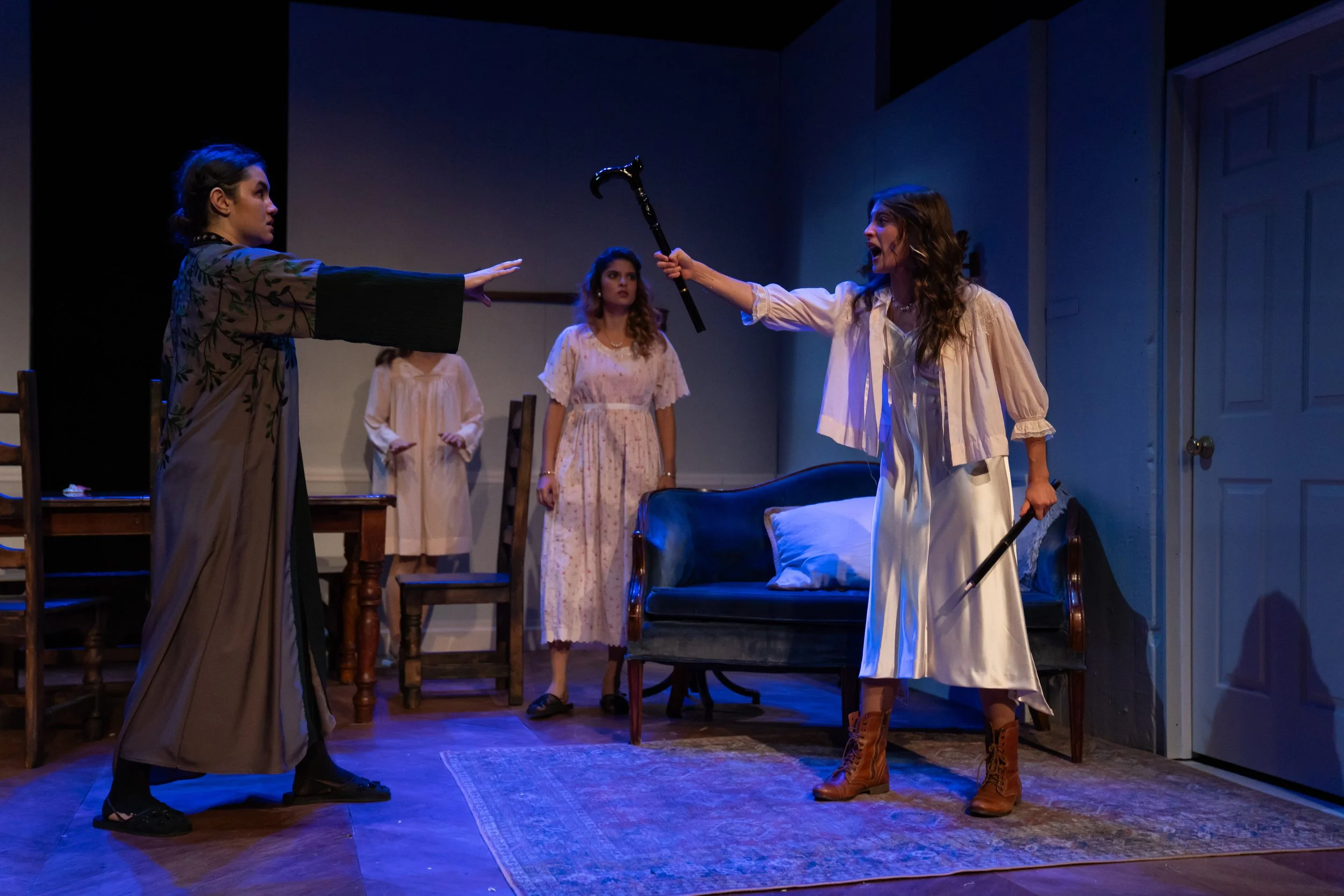 Four women in vintage nightgown styles are on a stage, engaged in a theatrical performance. One woman with a cane and white nightgown appears to be shouting or arguing with another woman wearing a patterned dress and black boots, who is extending her