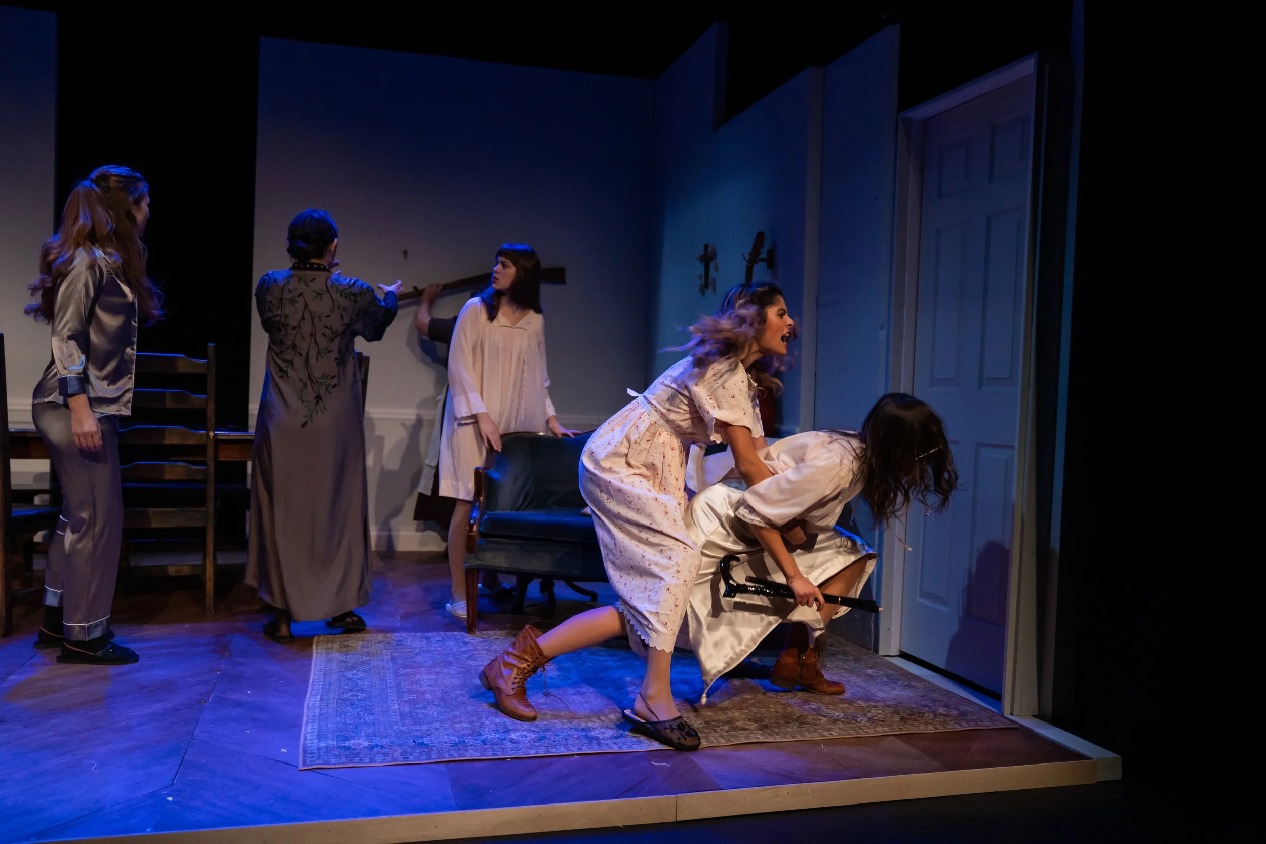 Scene from a theatrical play with multiple women in vintage pajamas and robes, some sitting or kneeling on a rug, others standing, inside a room with a closed door and decorative wall sconces, during an emotional moment.