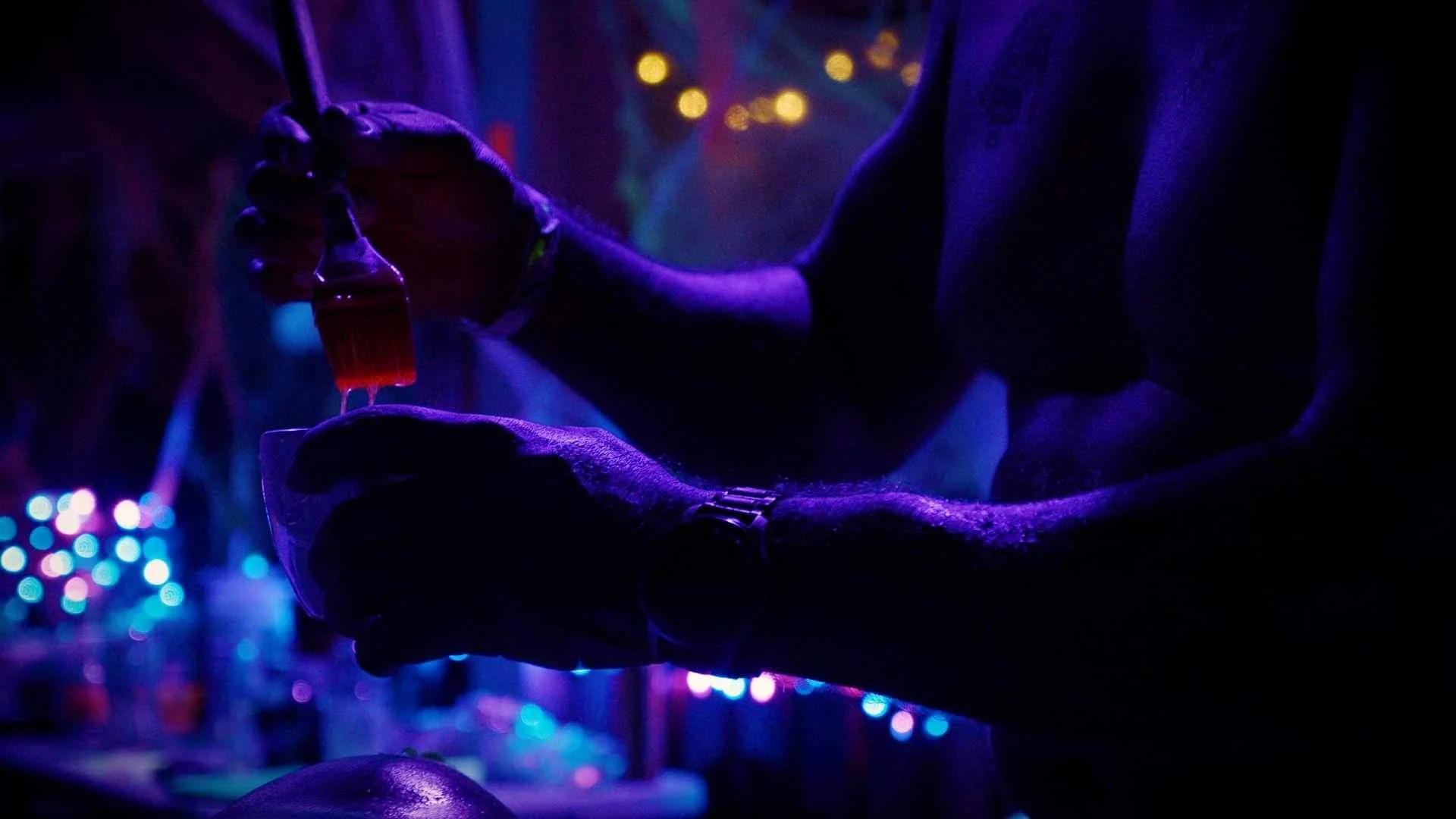 Person pouring red liquid from a beaker into a small cup in dark, neon-lit environment.