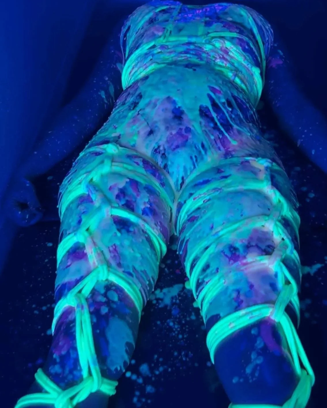 Person wearing glow-in-the-dark body paint and glowing leggings, lying on the floor in a dark environment with a black background.