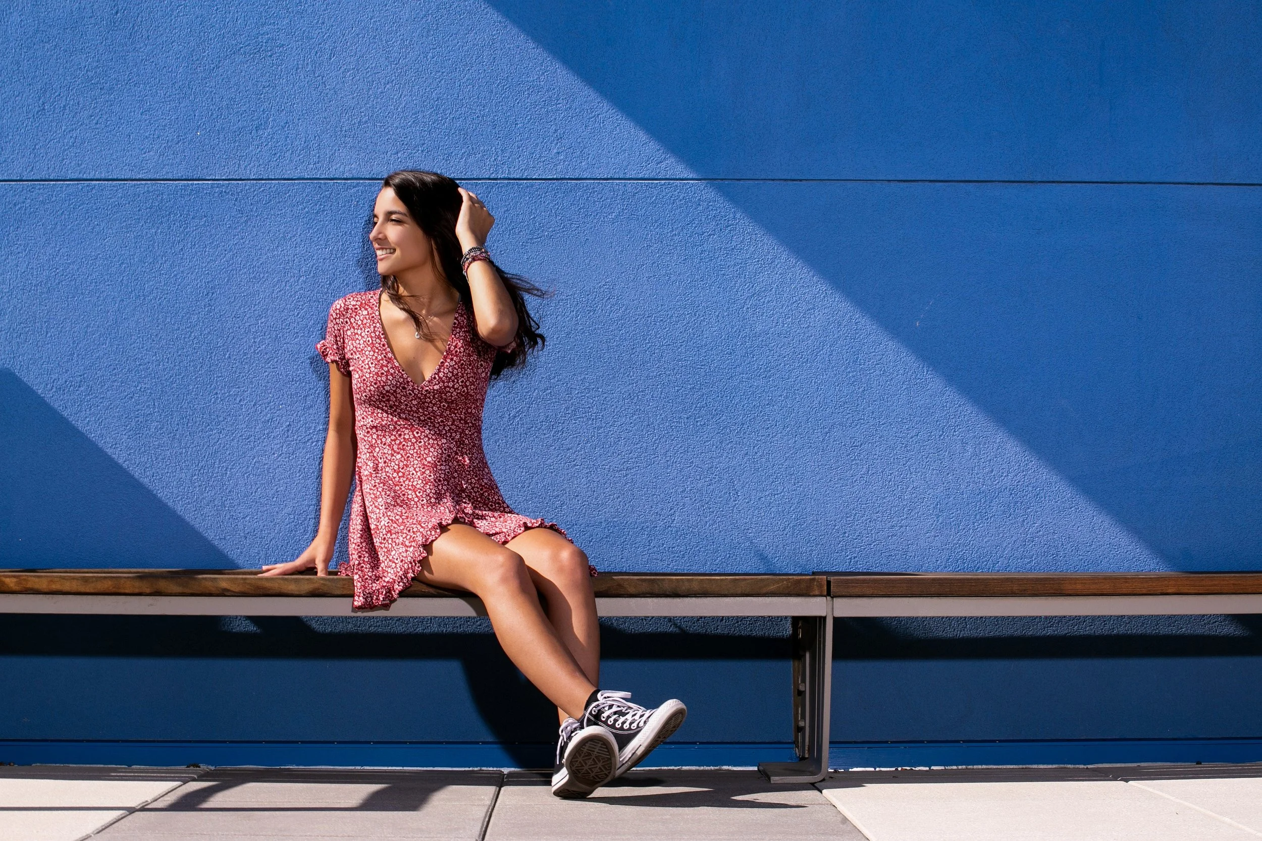 A young woman sitting on a wooden bench against a textured blue wall, smiling and looking to the left. She is wearing a red floral dress and black high-top sneakers, with her right hand touching her hair.