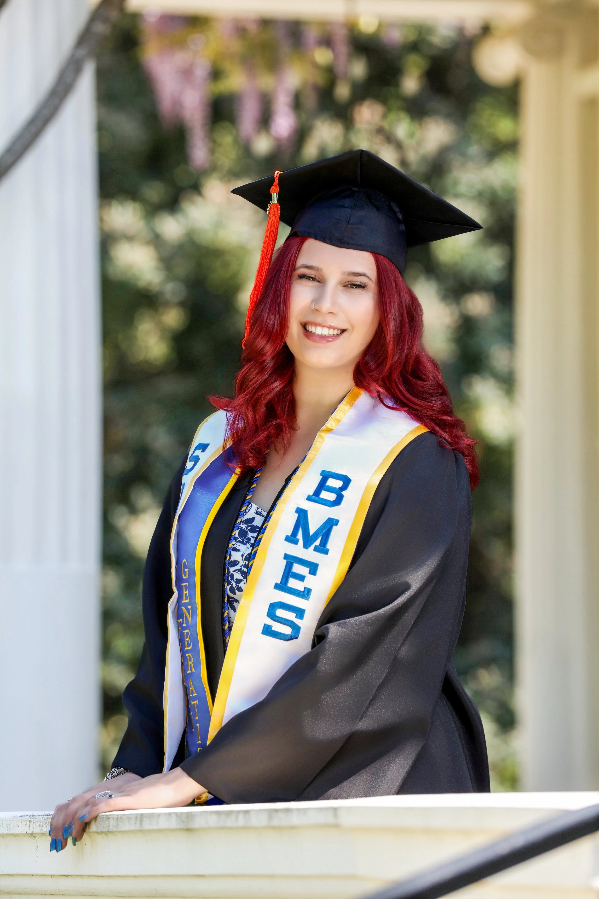 A woman with red hair wearing a black graduation cap and gown, standing outdoors with blurred trees in the background, displaying a white and blue honor stole.