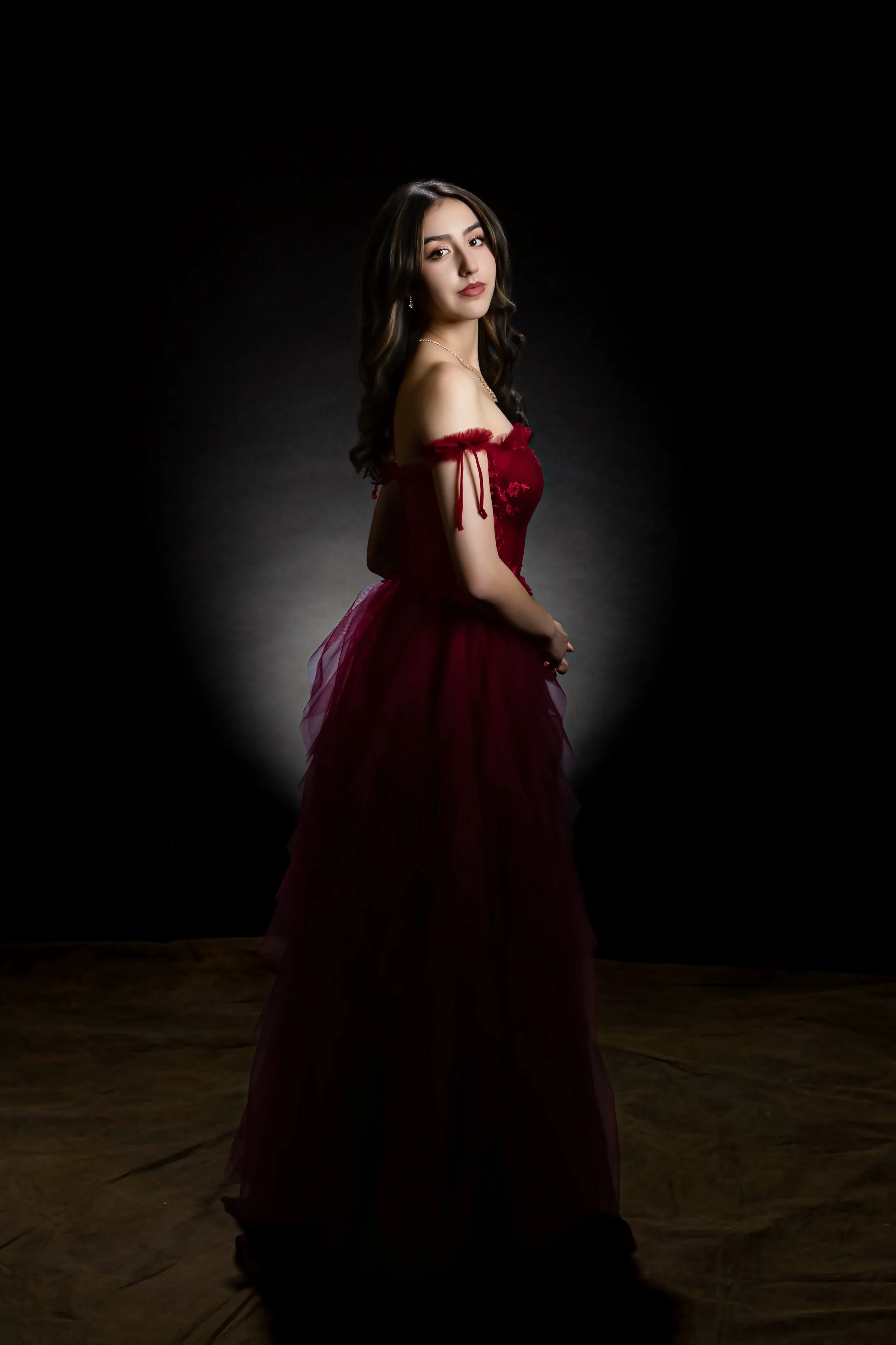 A young woman with long dark wavy hair wearing an off-shoulder red evening gown with lace details and a tulle skirt standing on a dark stage with a spotlight focused on her.