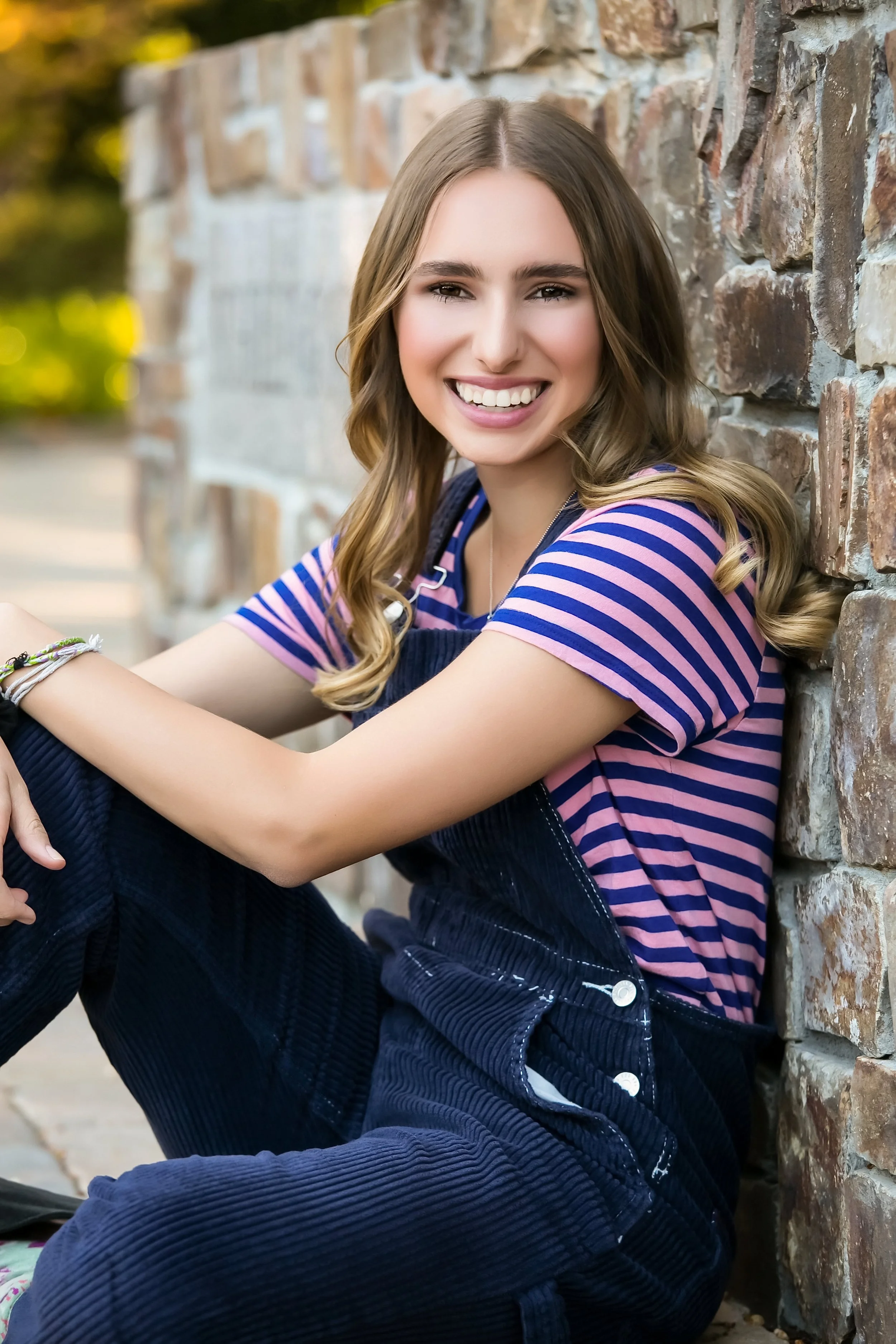 Young woman with long, wavy brown hair, smiling, sitting against a brick wall outdoors, wearing a pink and blue striped shirt and dark overalls.