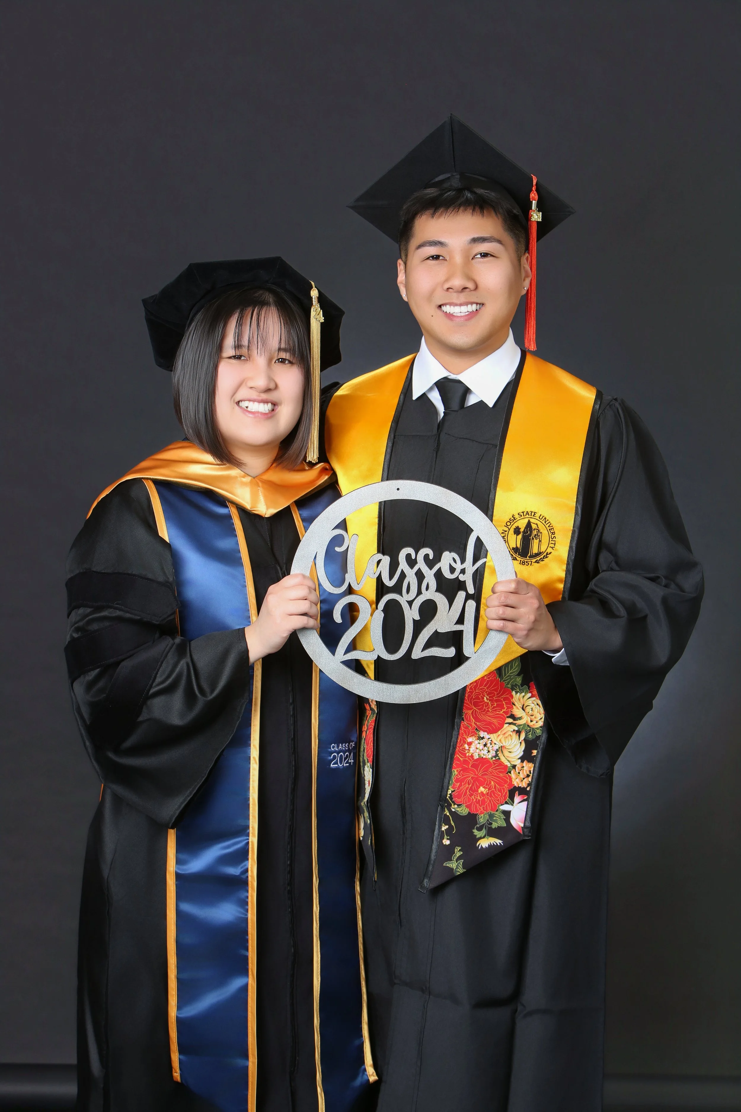 Two students in graduation caps and gowns holding a sign that reads "Class of 2024" in front of a dark background.