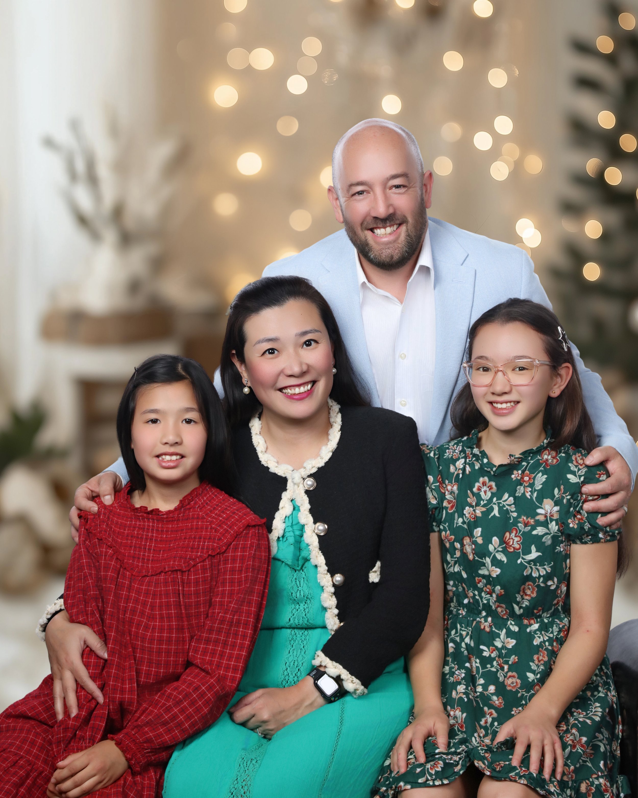 A group of people, including children and adults, celebrating indoors with festive Christmas lights in the background. They are smiling and dressed in colorful, holiday-themed clothing.
