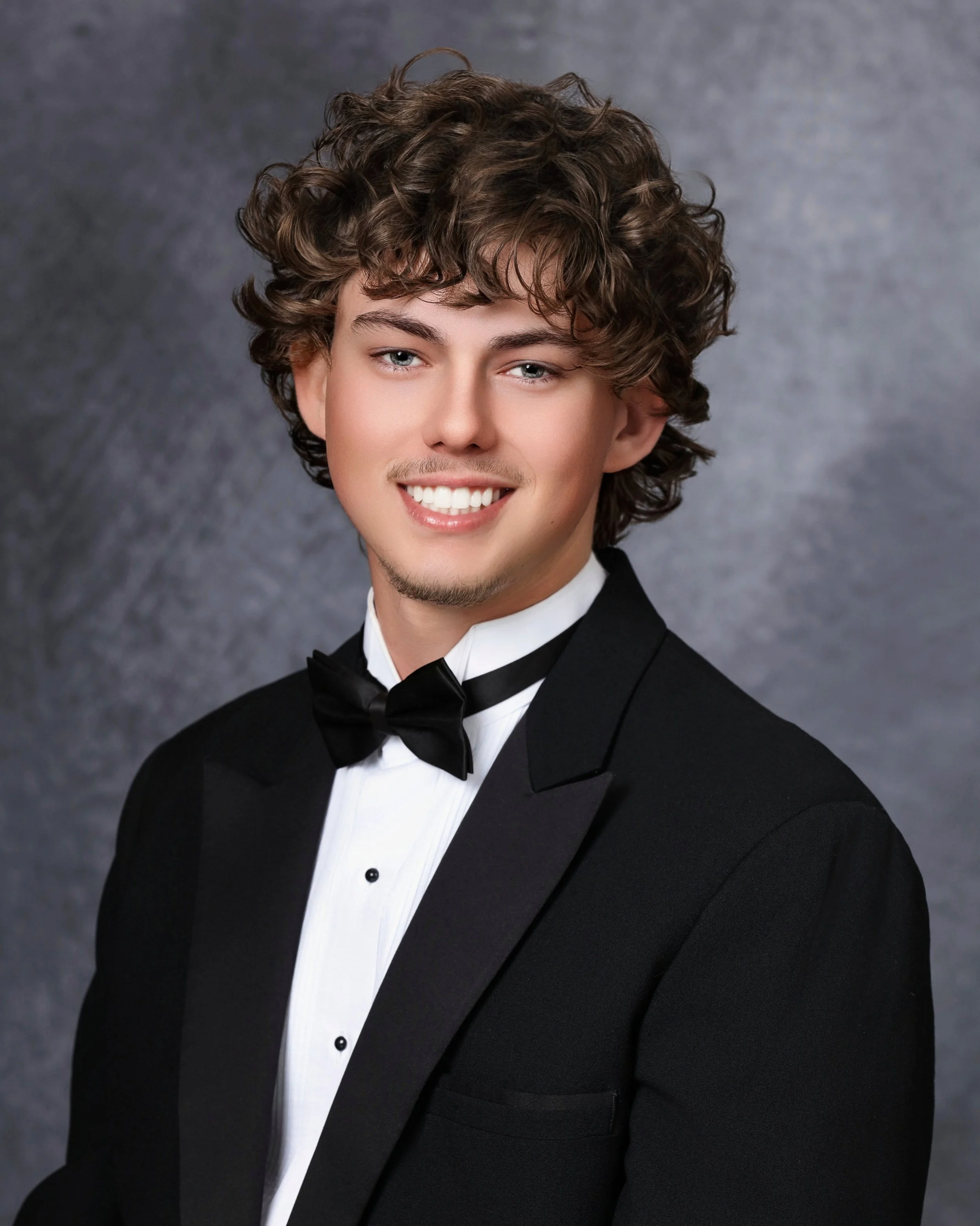 Young man with curly brown hair, blue eyes, and a light complexion, dressed in a tuxedo with a white shirt and black bow tie, smiling against a dark, textured background.
