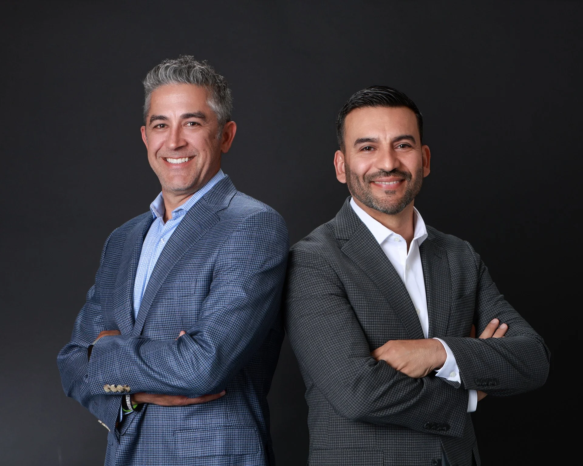Two men wearing suits standing against a dark background, smiling with crossed arms.