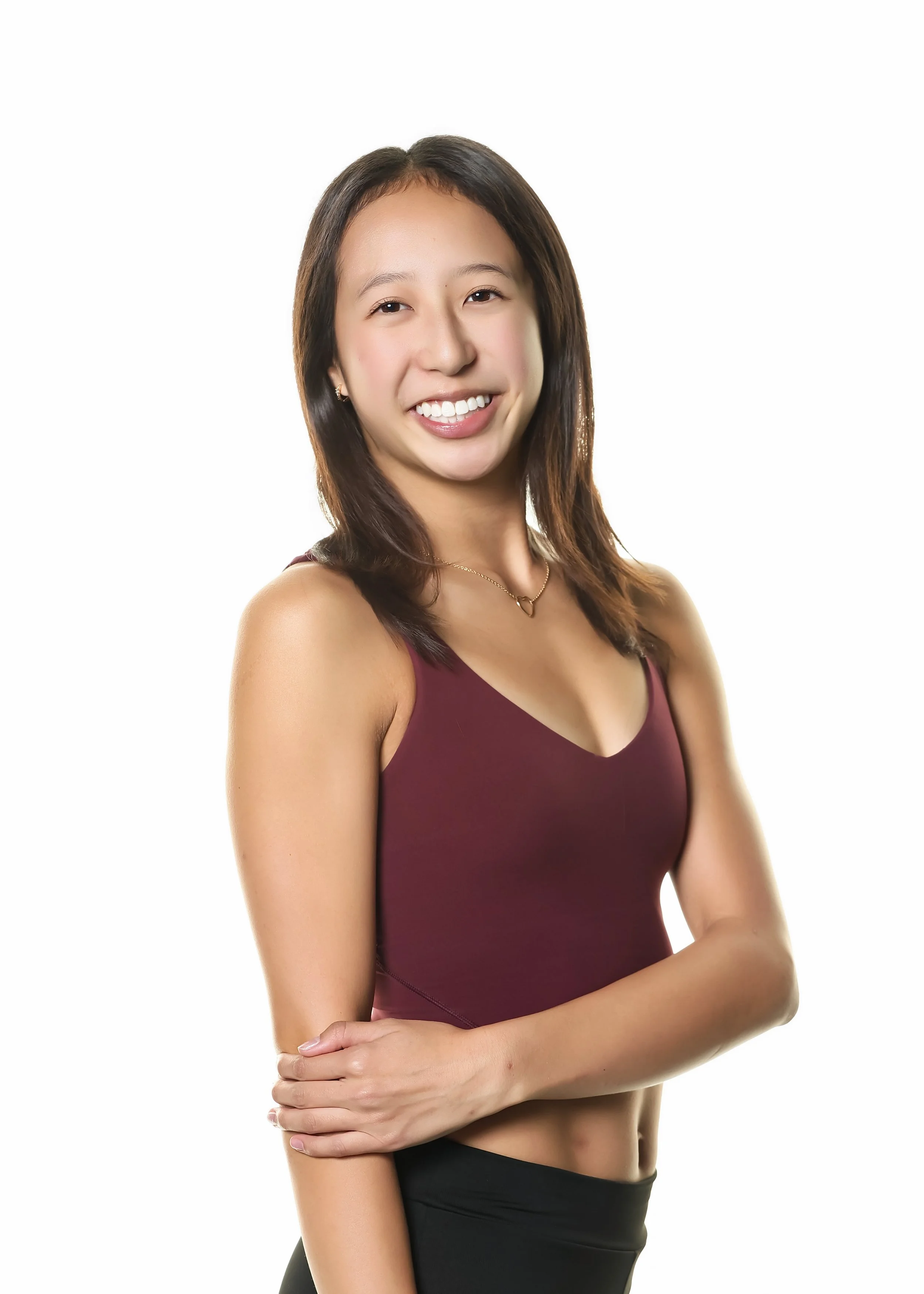 A young woman with long dark hair, smiling, wearing a sleeveless maroon top and a dainty necklace, standing against a white background.