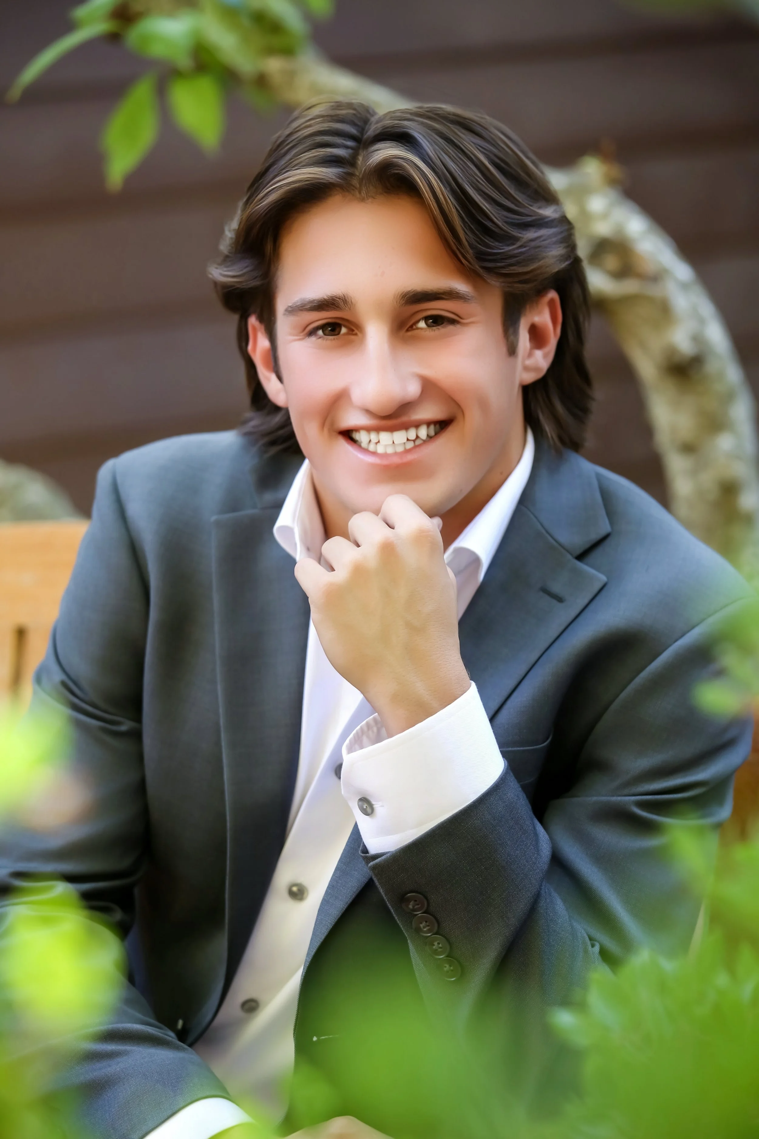 high school senior male wearing gray blazer posed outdoors on bench surrounded by plants.