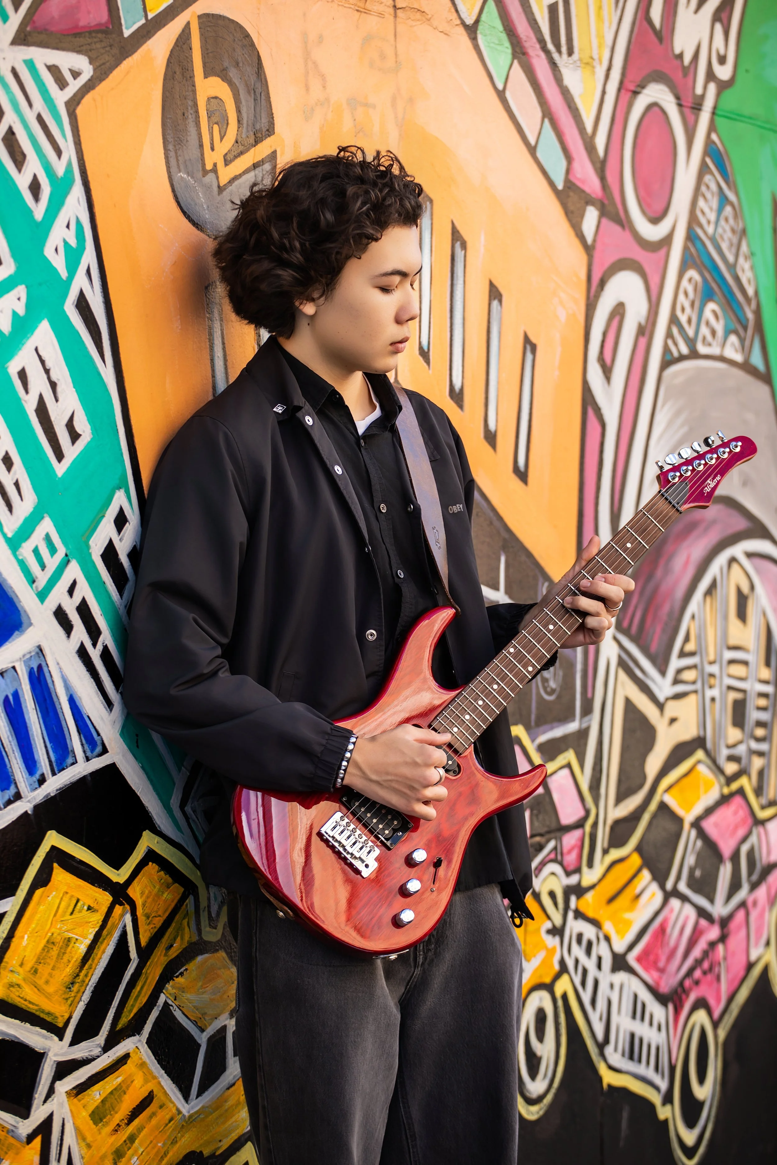 A young person with curly hair playing a red electric guitar against a colorful graffiti wall.