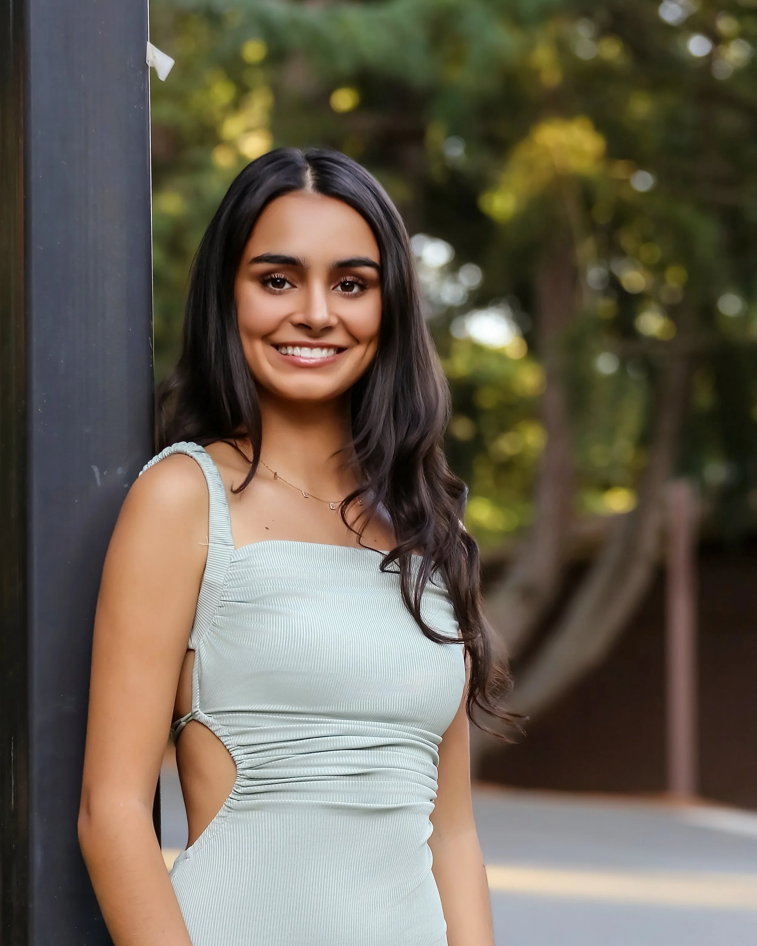 A young woman with long, dark hair smiling and standing outdoors, leaning against a black pole with a blurry green background.