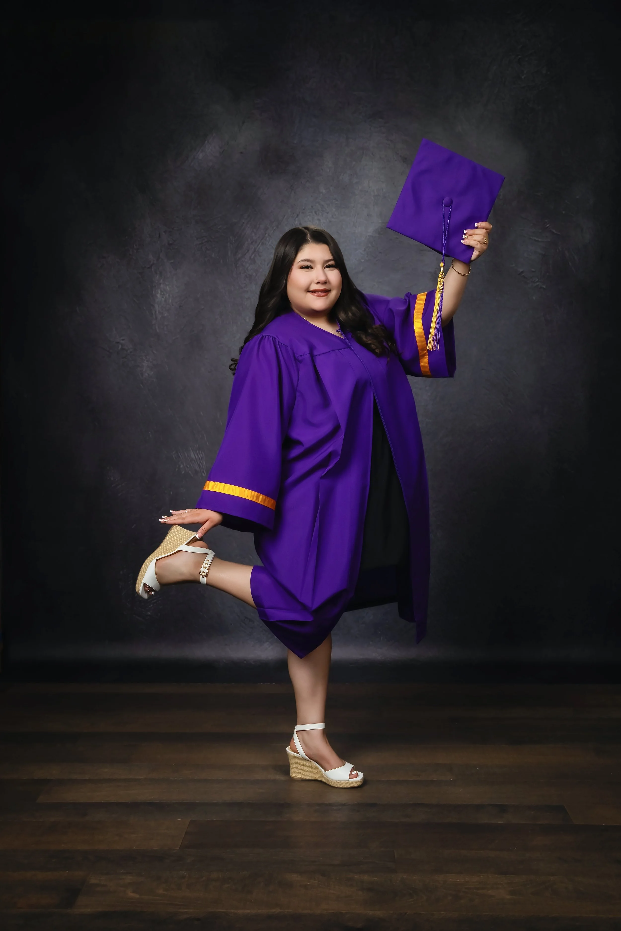 A woman in a purple graduation gown and cap, holding a diploma, posing with one leg bent back and wearing white wedge heels, standing on a wooden floor against a dark textured background.