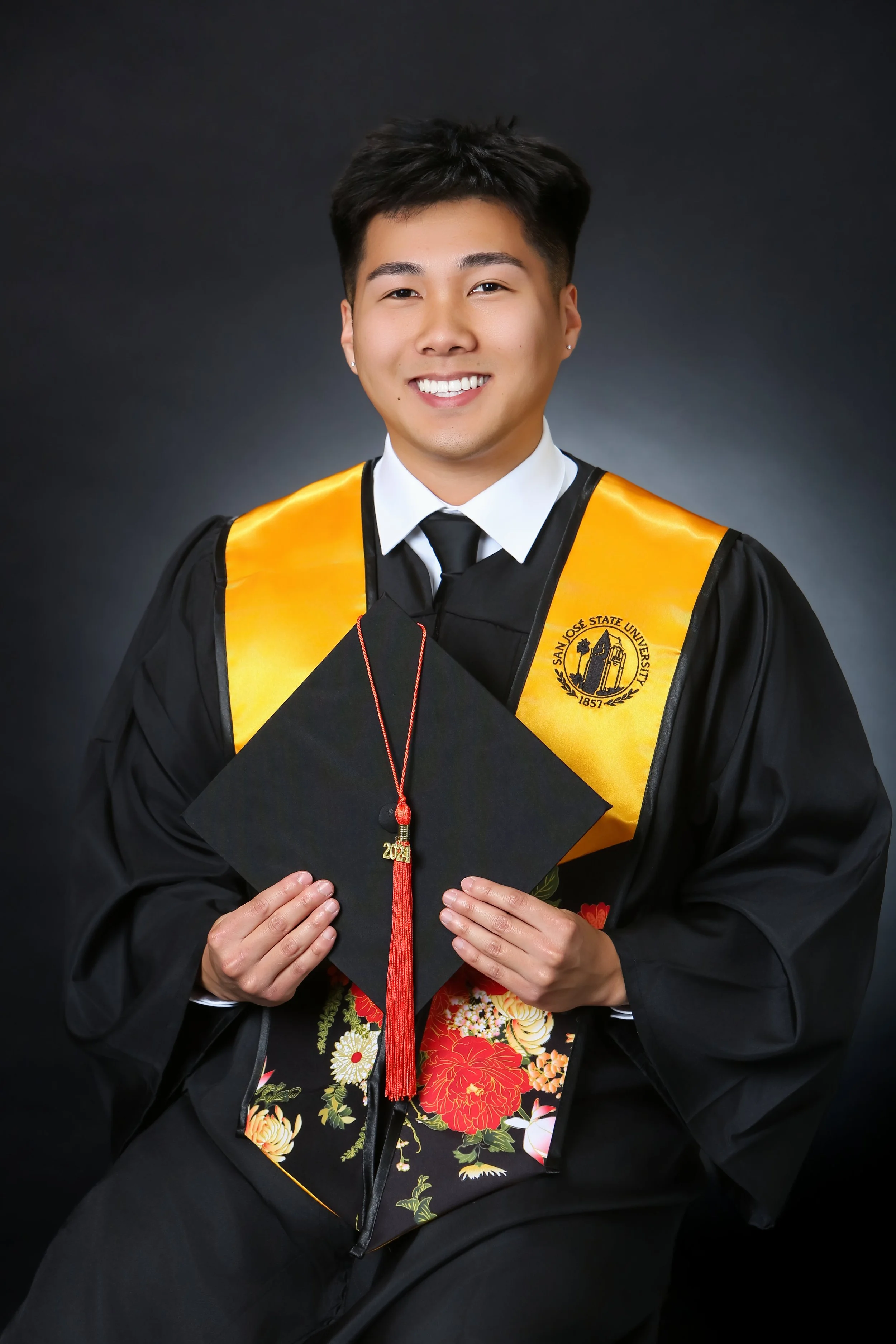 A young man in a graduation cap and gown holding a graduation diploma wearing a yellow sash with San Jose State University emblem.