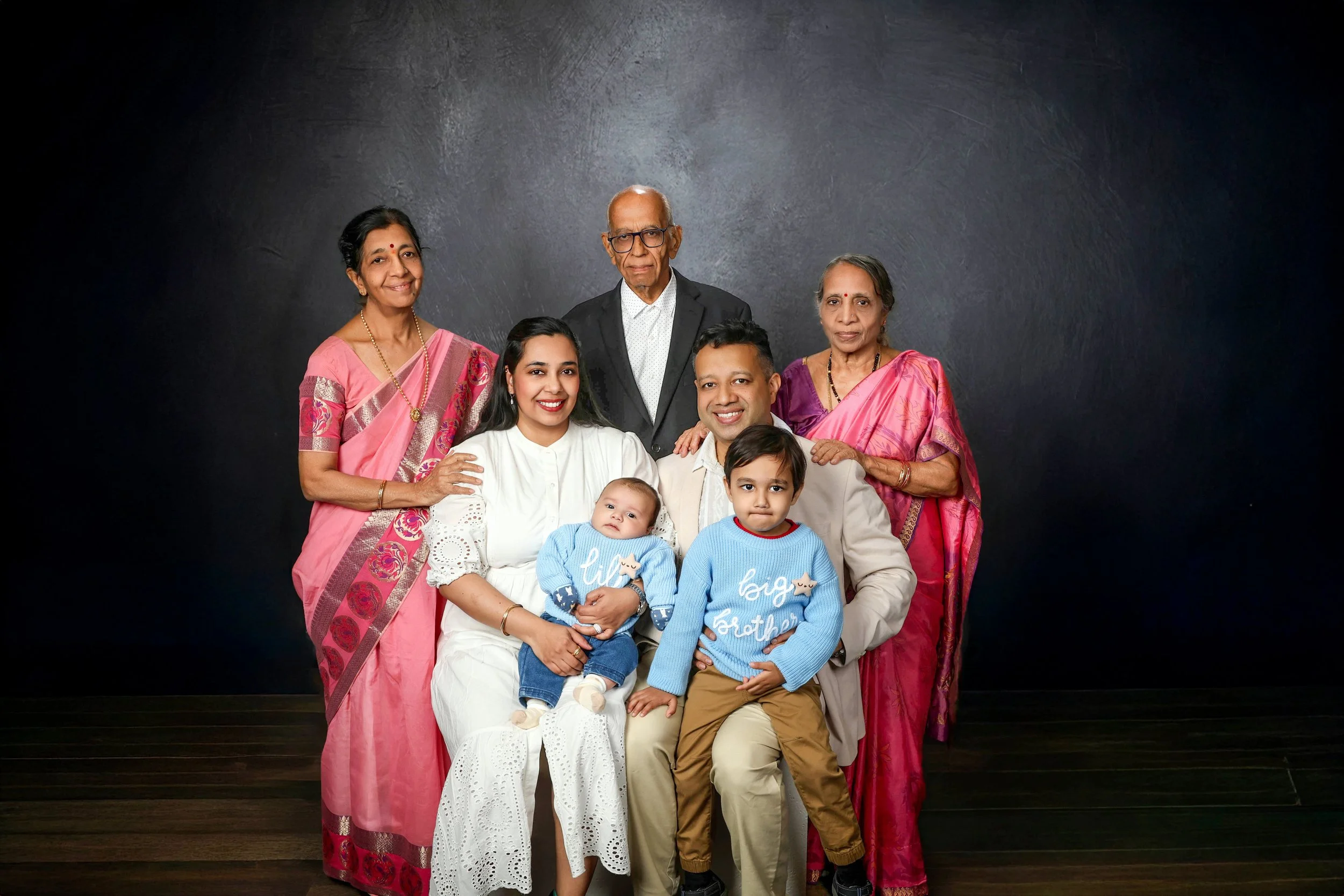 A multigenerational family portrait of nine people, including grandparents, parents, children, and babies, dressed in traditional and modern clothing, standing against a dark backdrop in an indoor setting.