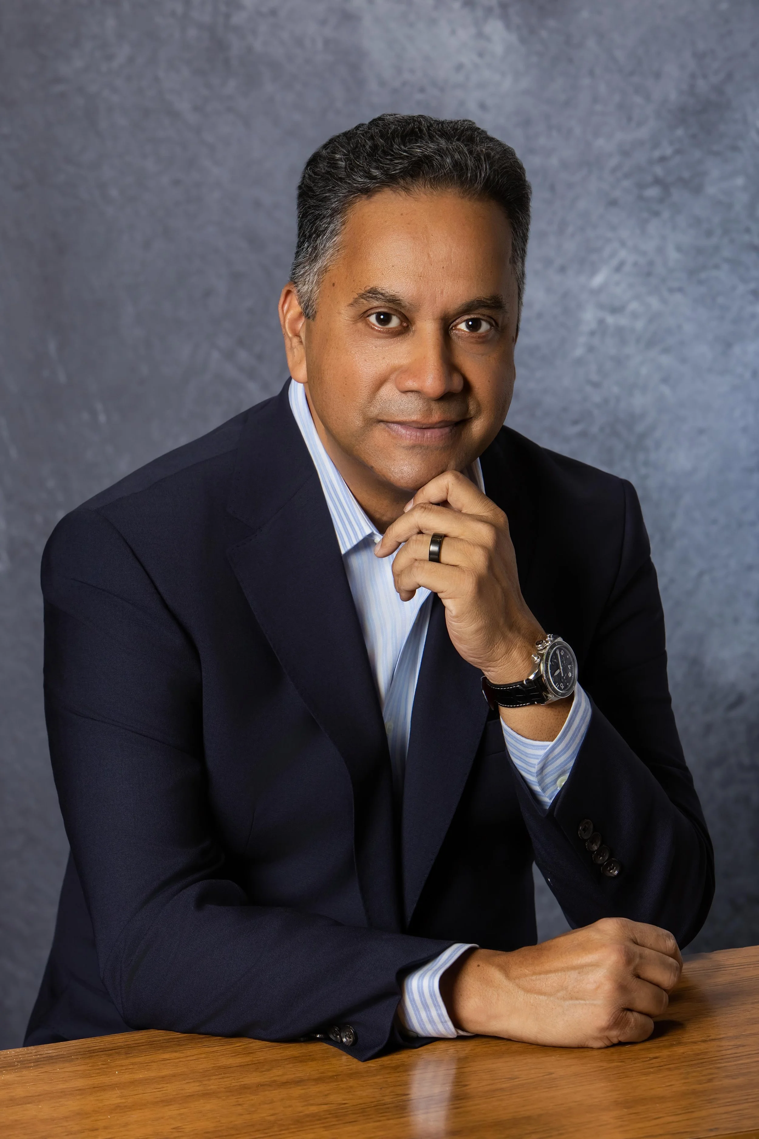 A man in a suit sitting at a wooden desk, with his right hand resting on his chin and his left hand on the table, against a gray textured background.