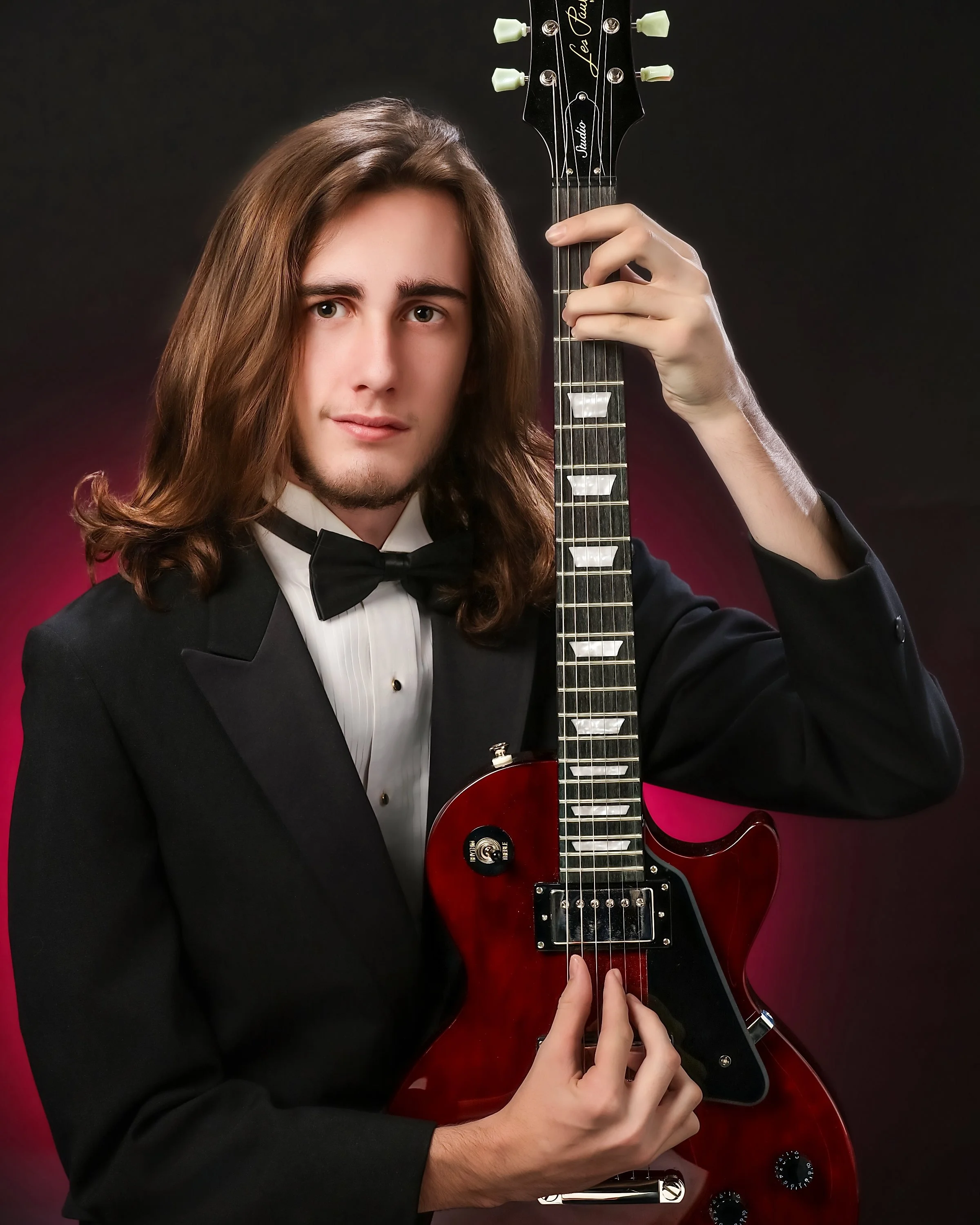 A young man with long brown hair wearing a tuxedo and bowtie, holding a red electric guitar and playing it with his left hand on the fretboard and his right hand on the strings. The background is dark with a hint of pinkish-red light.