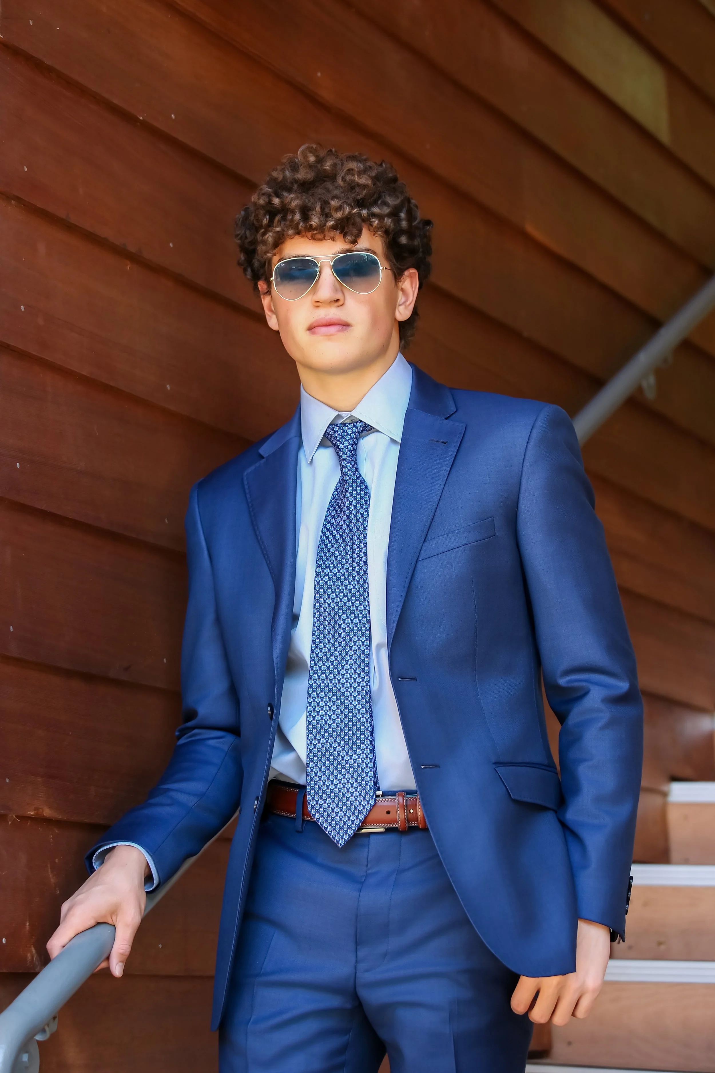 A young man with curly hair wearing a blue suit, tie, and sunglasses, leaning on a railing in front of a wooden wall.