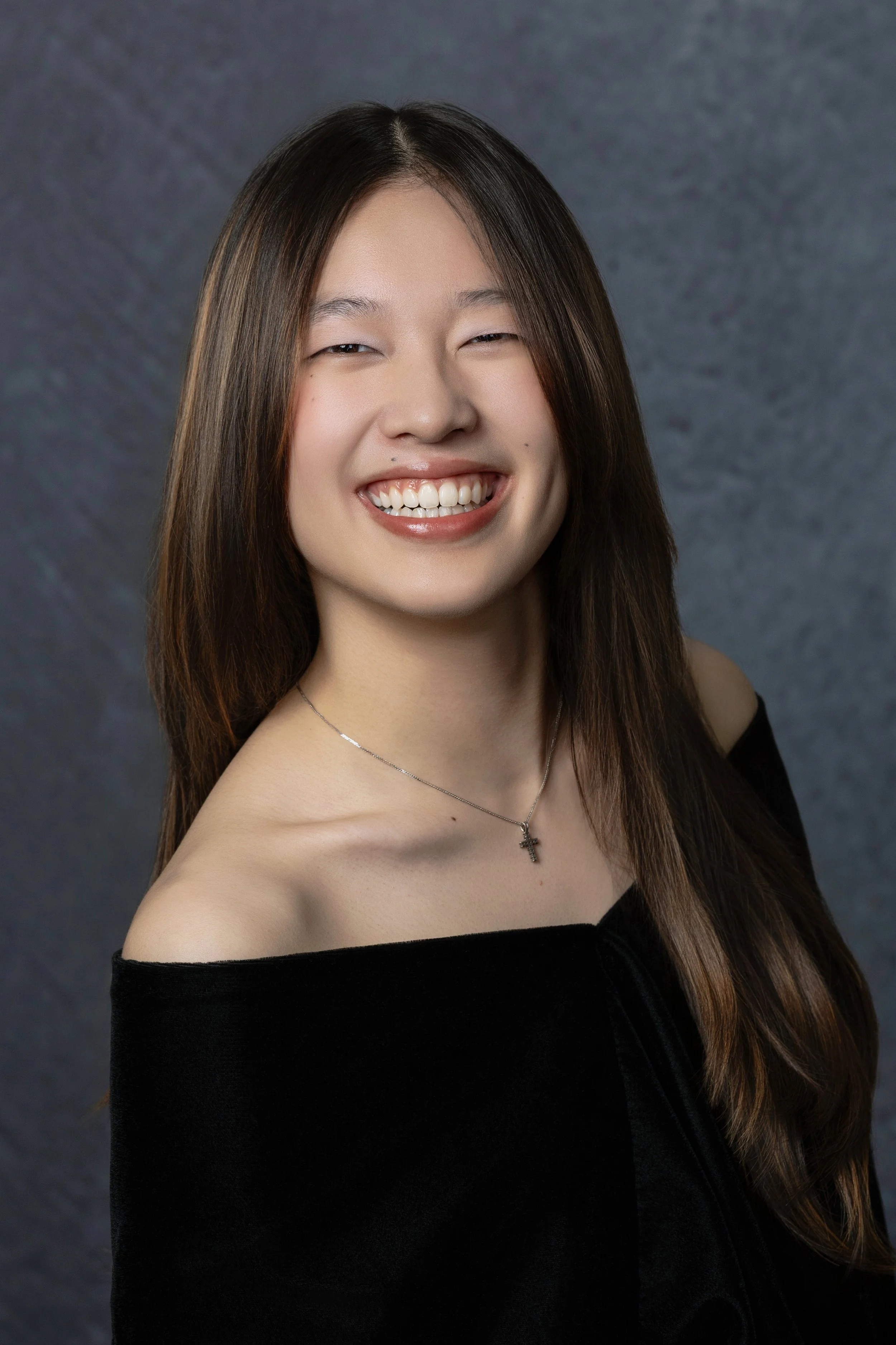 A young woman with long brown hair, smiling, wearing a black off-shoulder top, a silver necklace with a cross pendant, against a gray textured background.