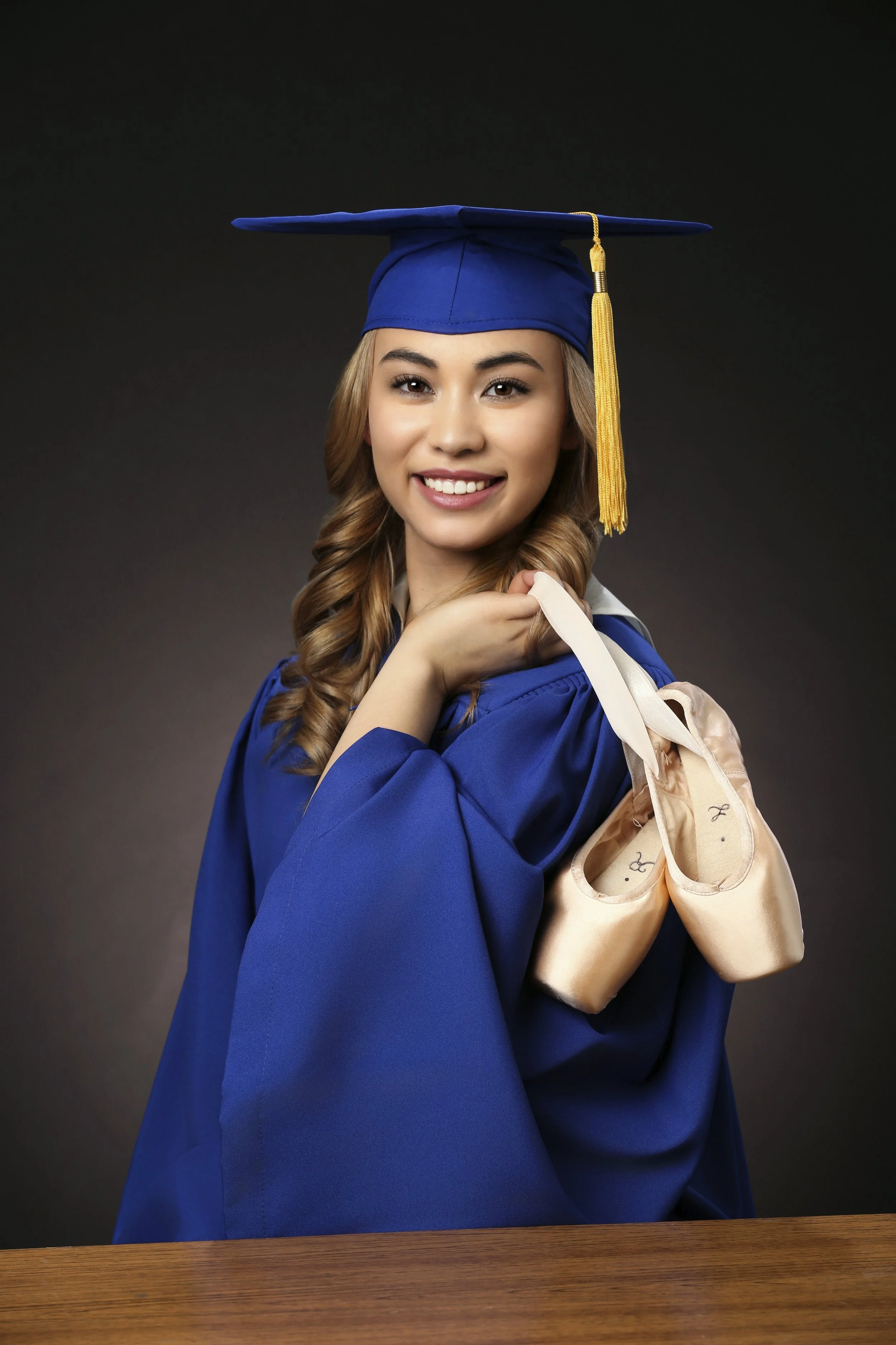 A young woman in a blue graduation cap and gown holding a pair of pink ballet shoes, standing against a dark background.