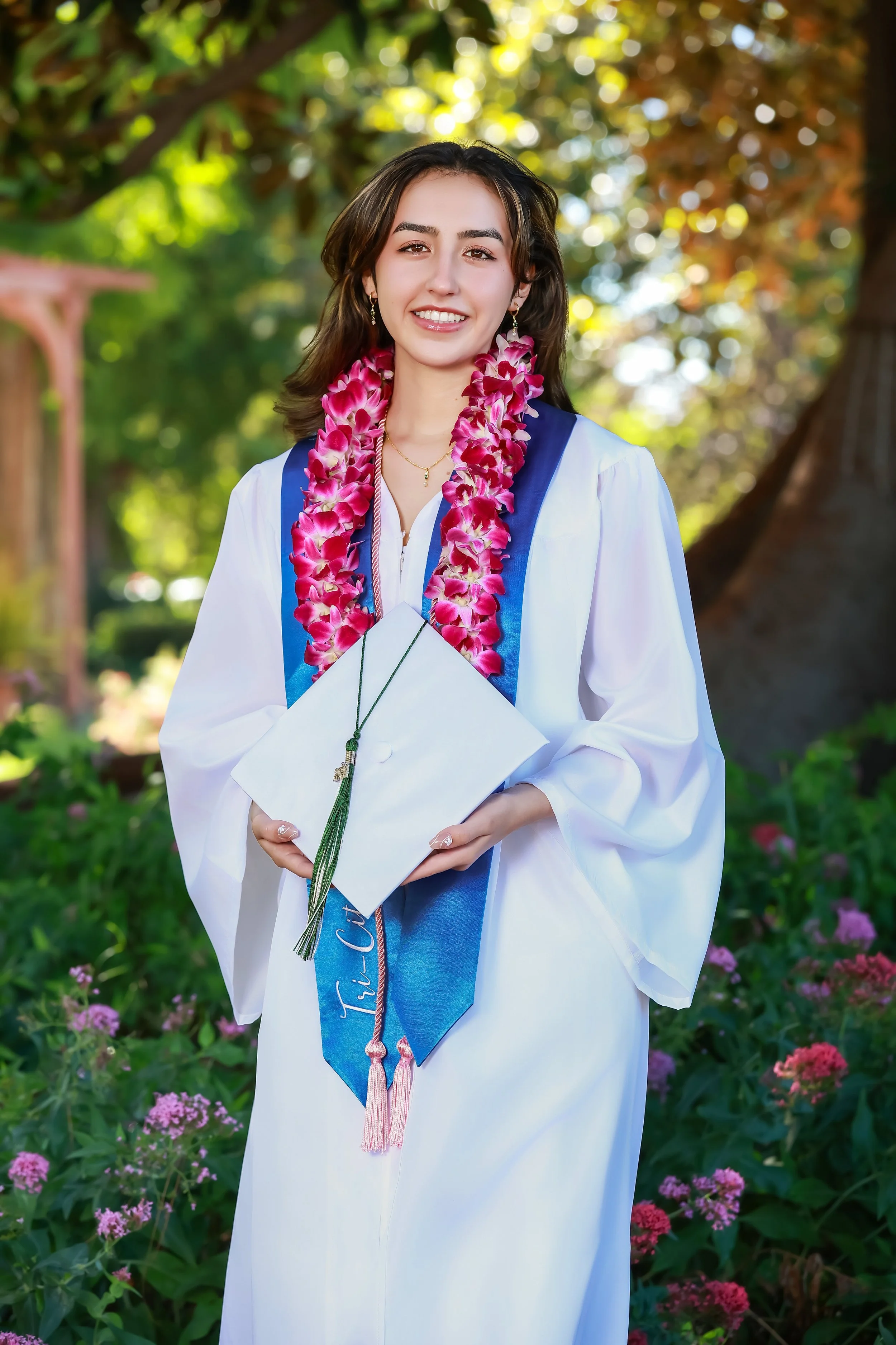 A young woman in a graduation gown holding a diploma, wearing a pink flower lei and smiling, standing outdoors with trees and pink flowers in the background.
