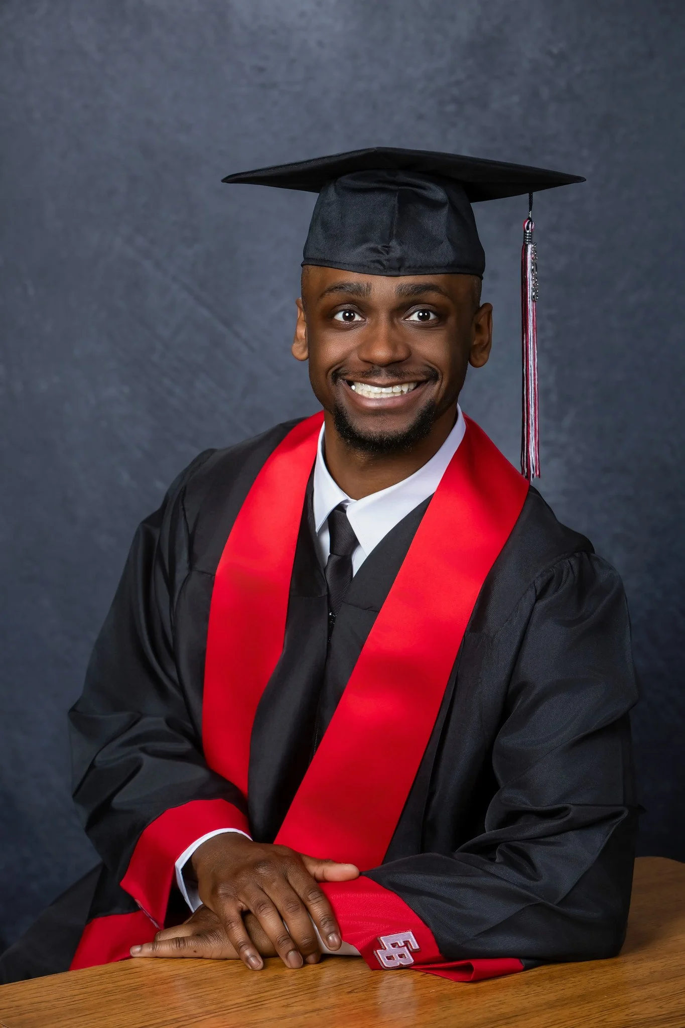 A young man in a graduation cap and gown with a red stole, smiling and sitting at a wooden table, against a gray background.