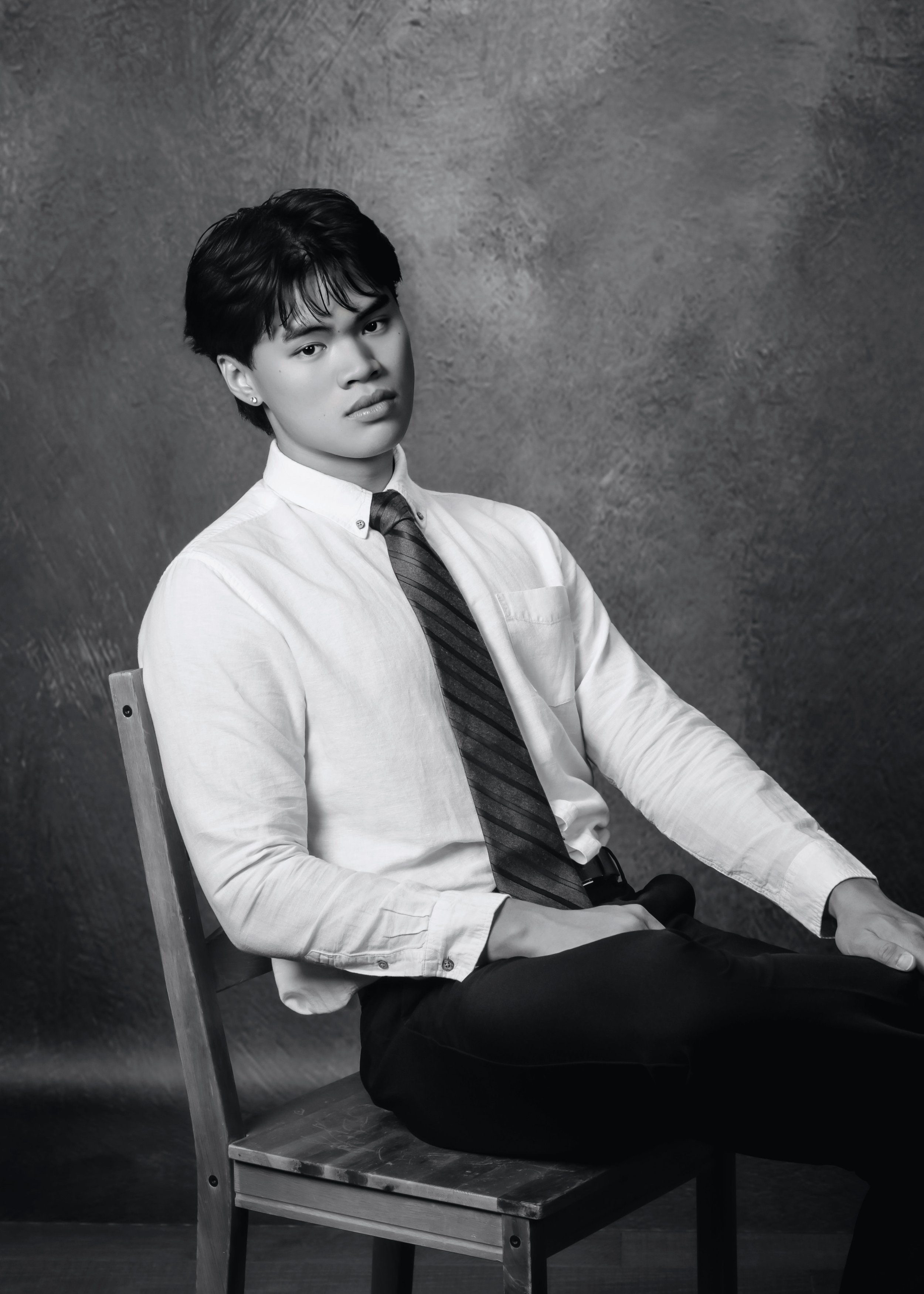Black and white portrait of a young man with dark hair, sitting on a wooden chair, wearing a white shirt, striped tie, and dark pants, against a textured wall background.