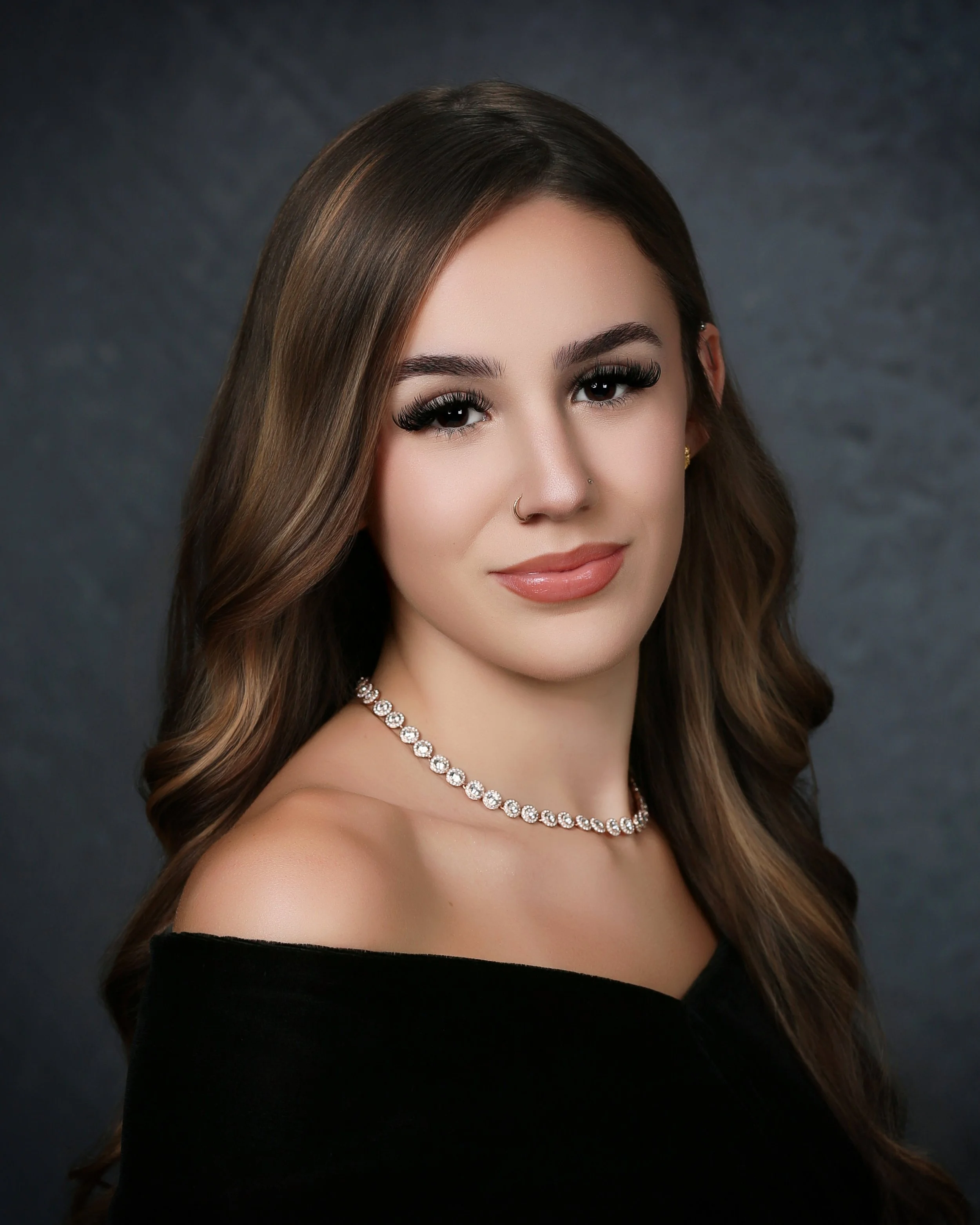 A woman with long, wavy brown hair and light makeup, wearing a diamond necklace, a small nose piercing, and a black off-the-shoulder top, smiling in front of a dark, blurred background.