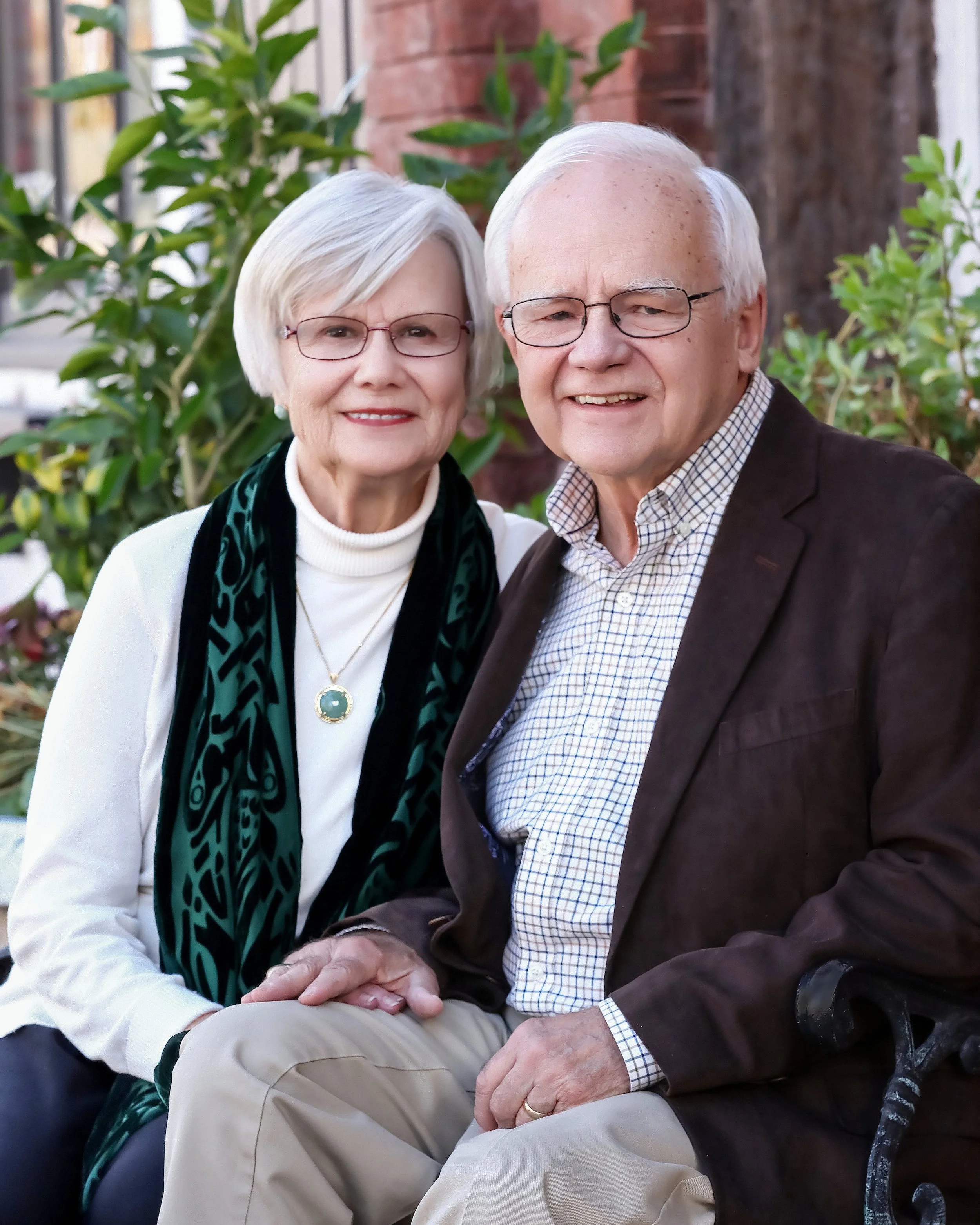An elderly couple sitting outdoors, holding hands, smiling, with greenery and a brick wall in the background.