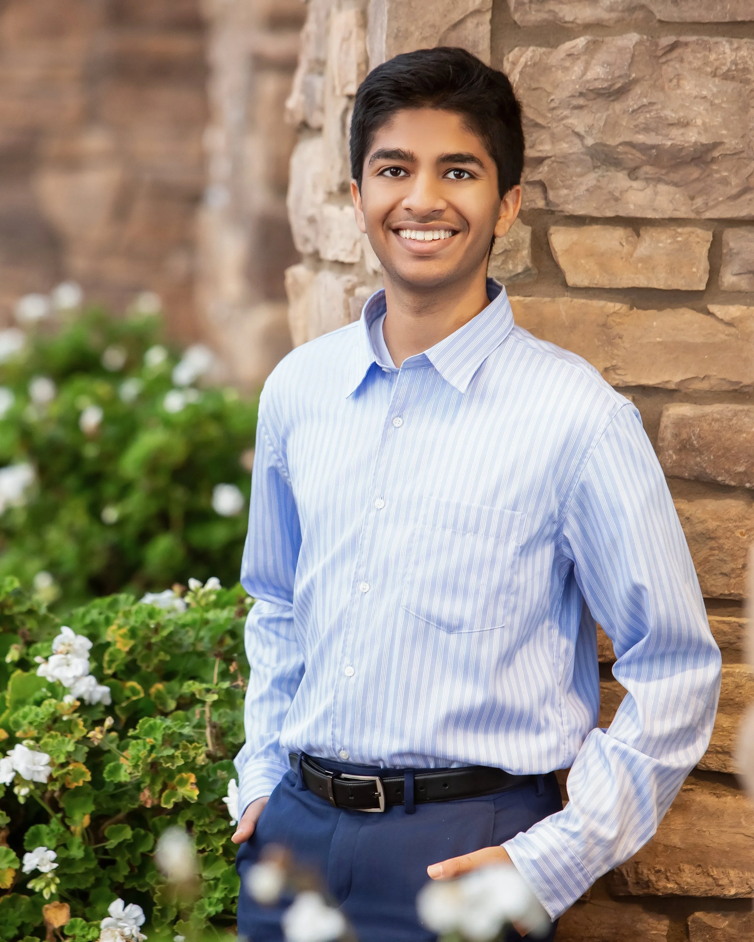 A young man with dark hair, wearing a blue striped dress shirt and navy pants with a black belt, standing outdoors against a brick wall and surrounded by green plants with white flowers, smiling at the camera.