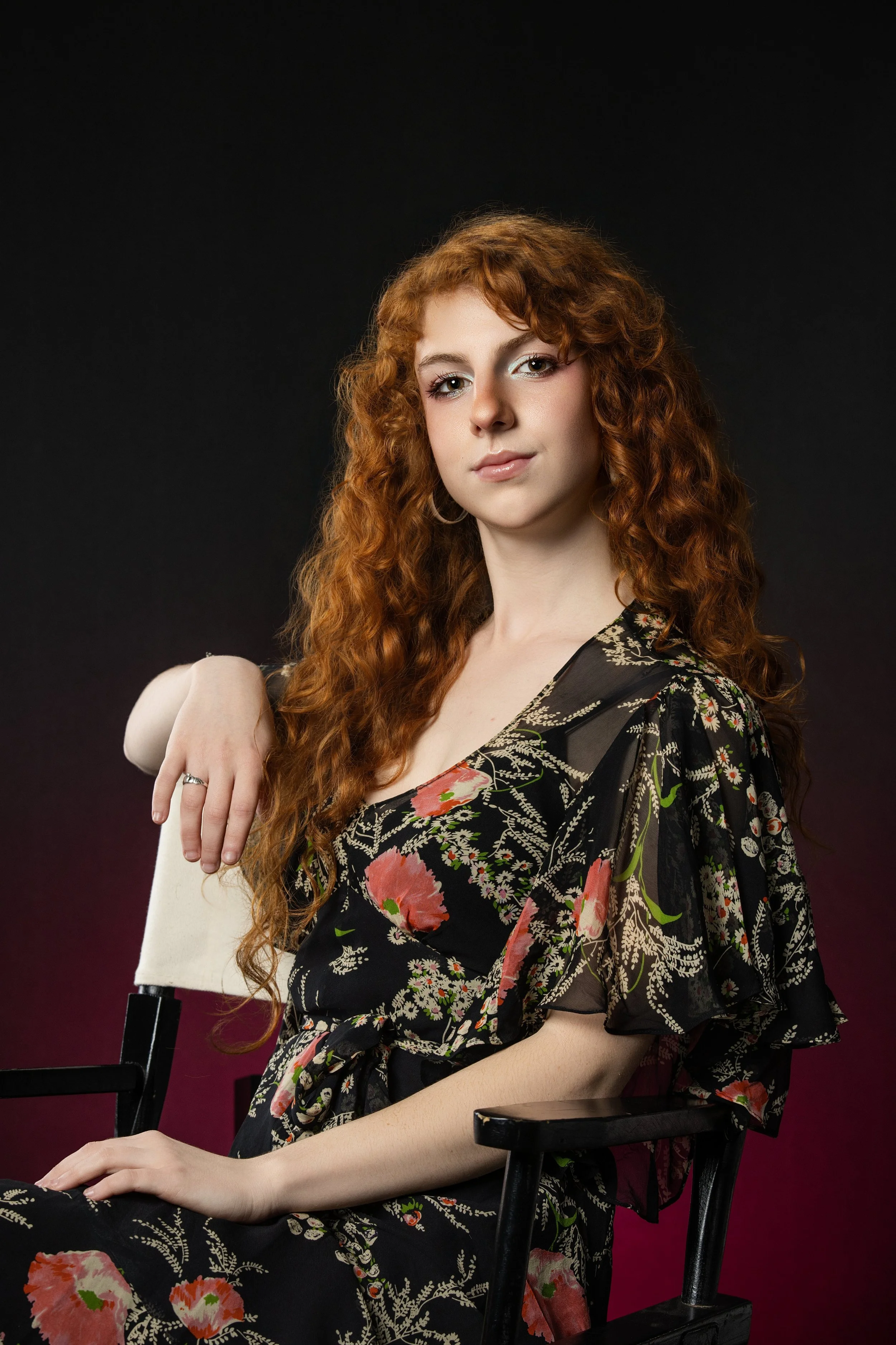 high school senior girl wearing a casual dress, sitting on a director's chair in studio with black and pink background