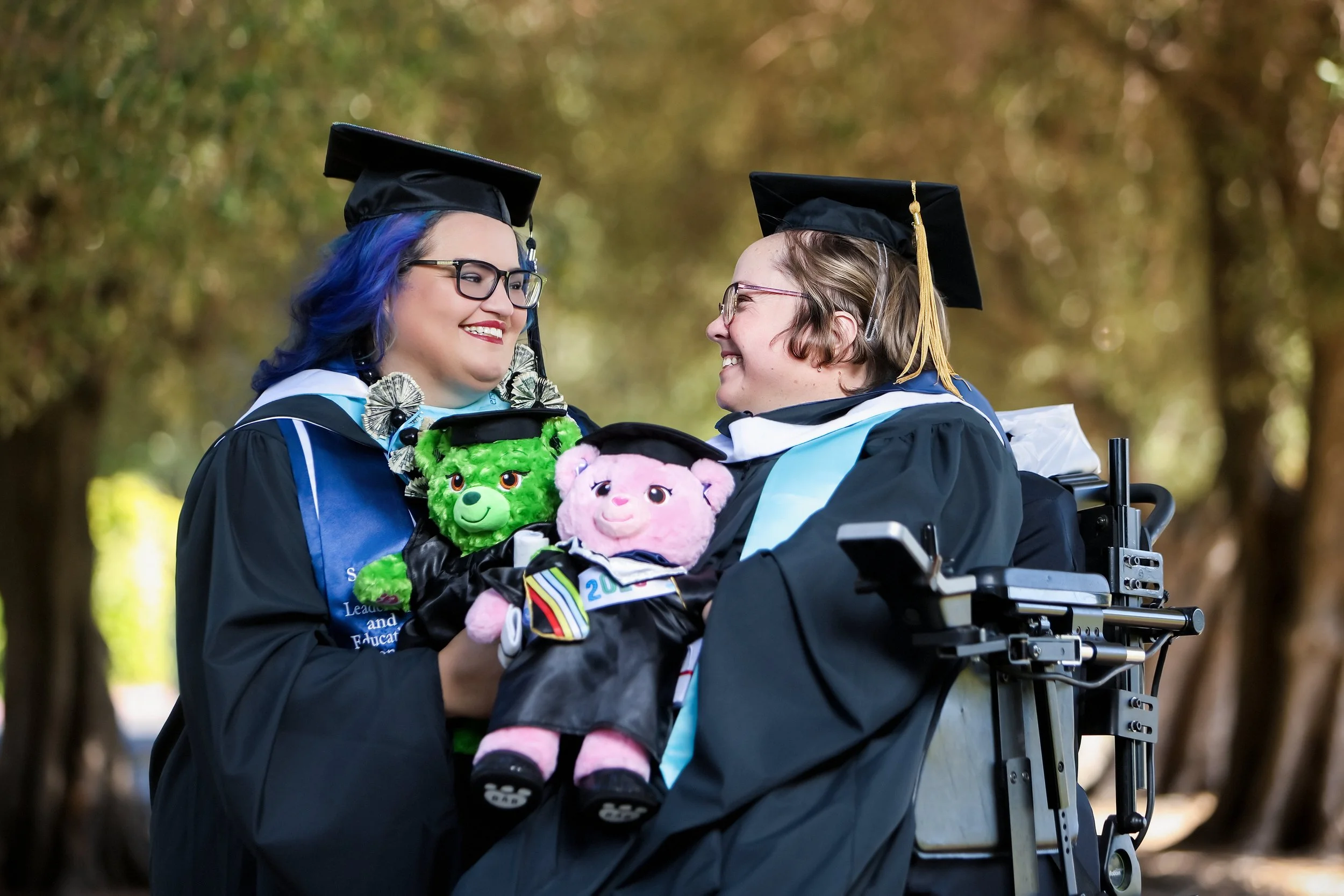 Two women in graduation caps and gowns smiling at each other outdoors, with trees in the background. One woman is in a wheelchair and holding stuffed animals.