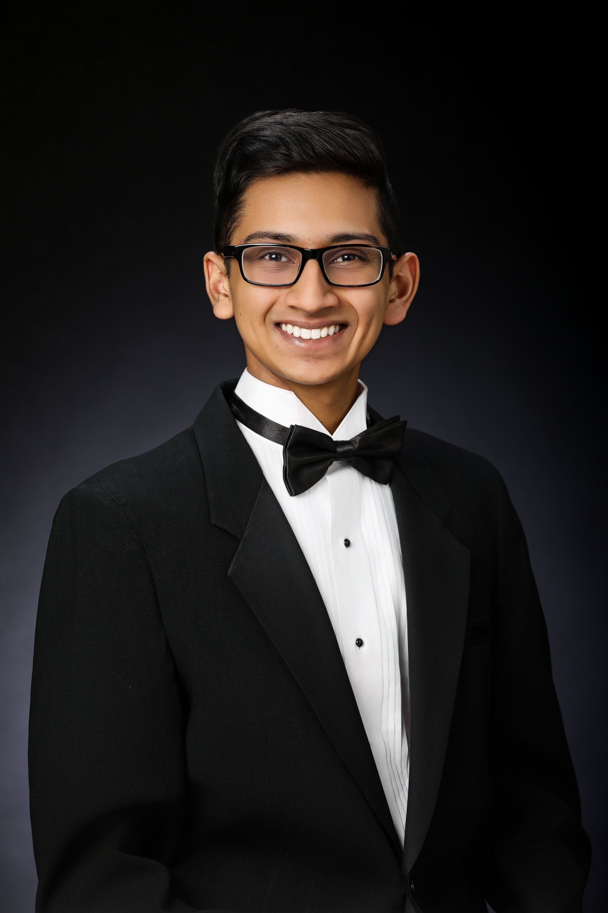Young man smiling, wearing black tuxedo and bow tie, glasses, with dark hair against a black background.