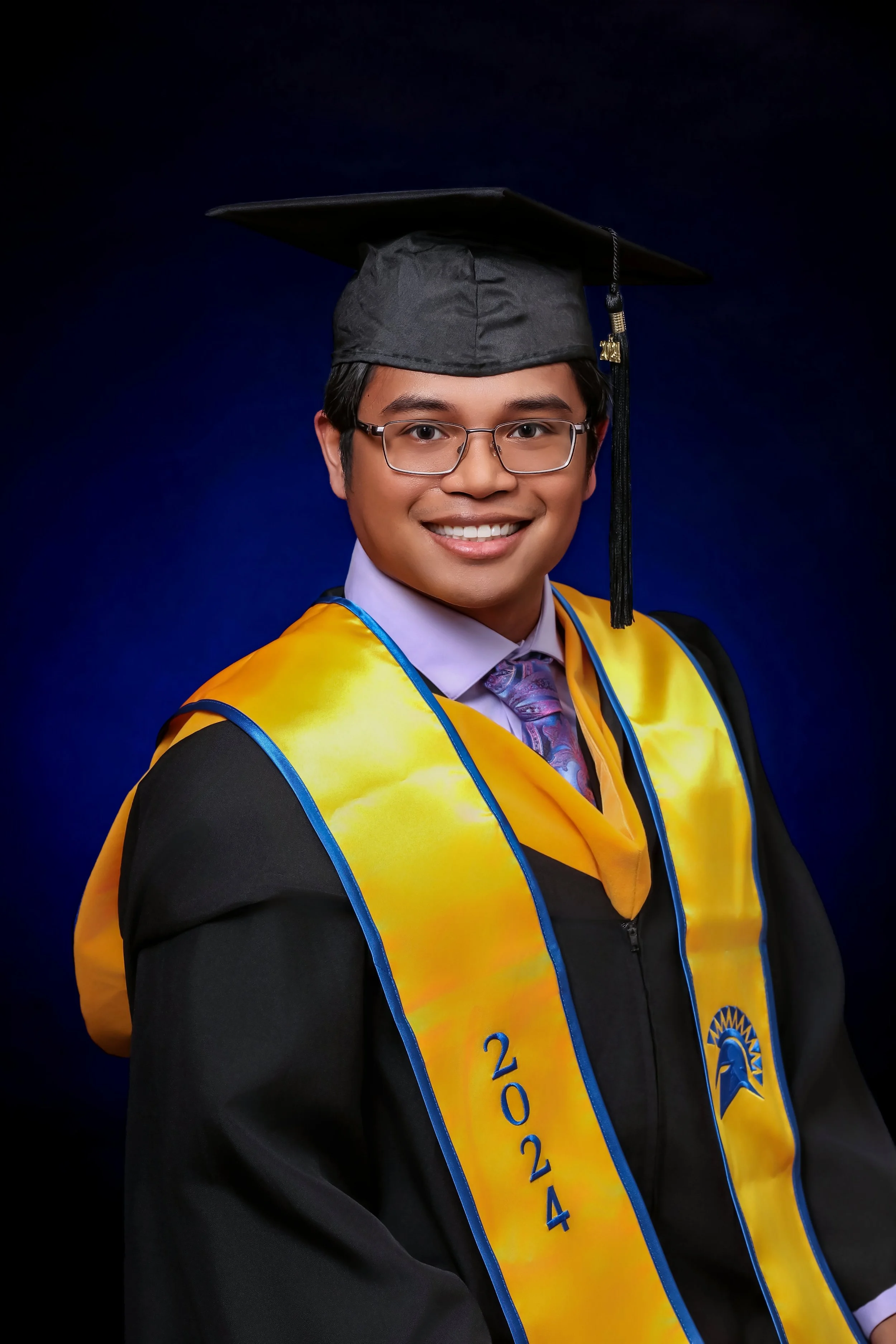A young man with glasses, dark hair, and a bright smile in a graduation gown and cap, wearing a yellow and blue stole that displays the year 2024, against a dark blue background.